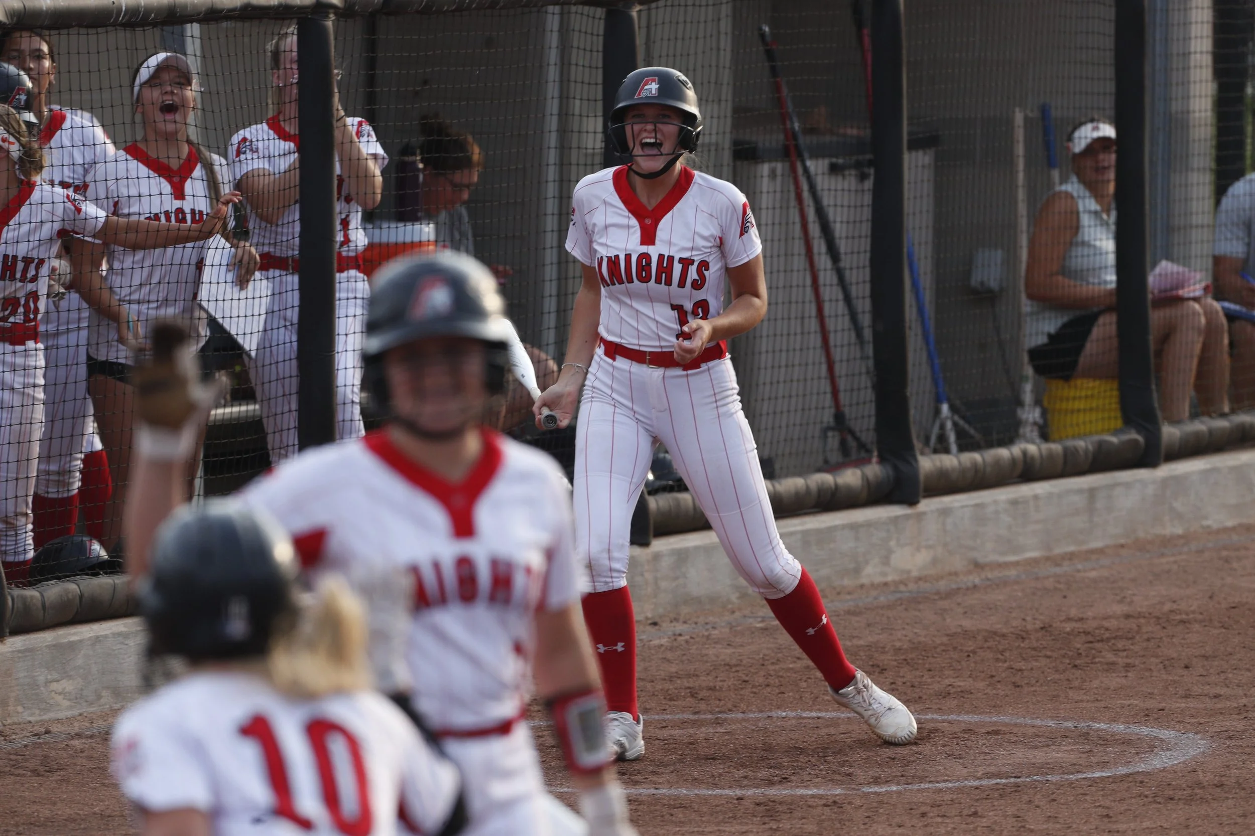 A softball player wearing a black helmet and white uniform with red accents, holding a bat, standing near the dugout fence, smiling. Other teammates and spectators are visible in the background.