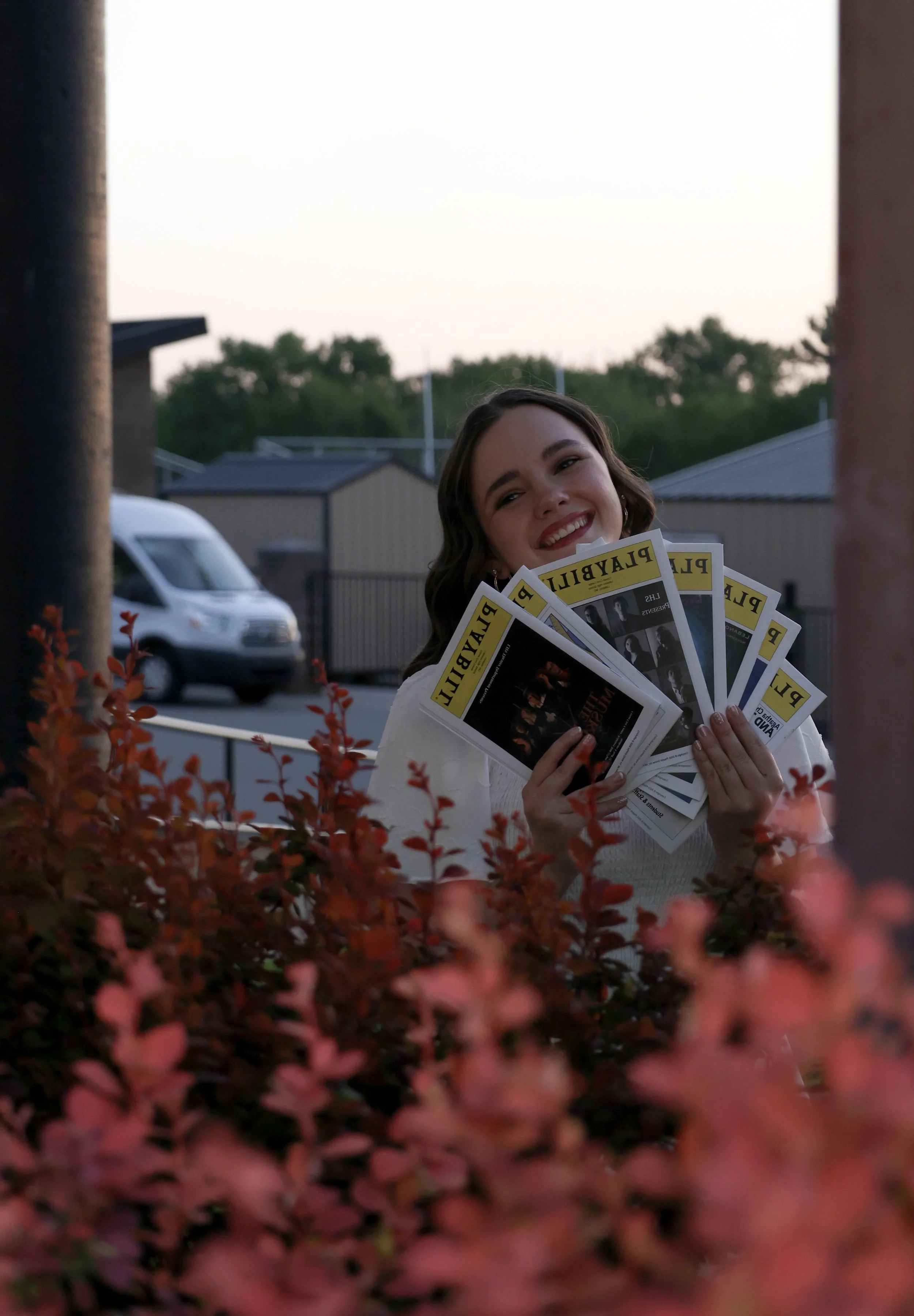 A smiling woman holding several Playbill programs in front of pinkish-red bushes outdoors with a small building, a parking lot, and trees in the background during daylight.
