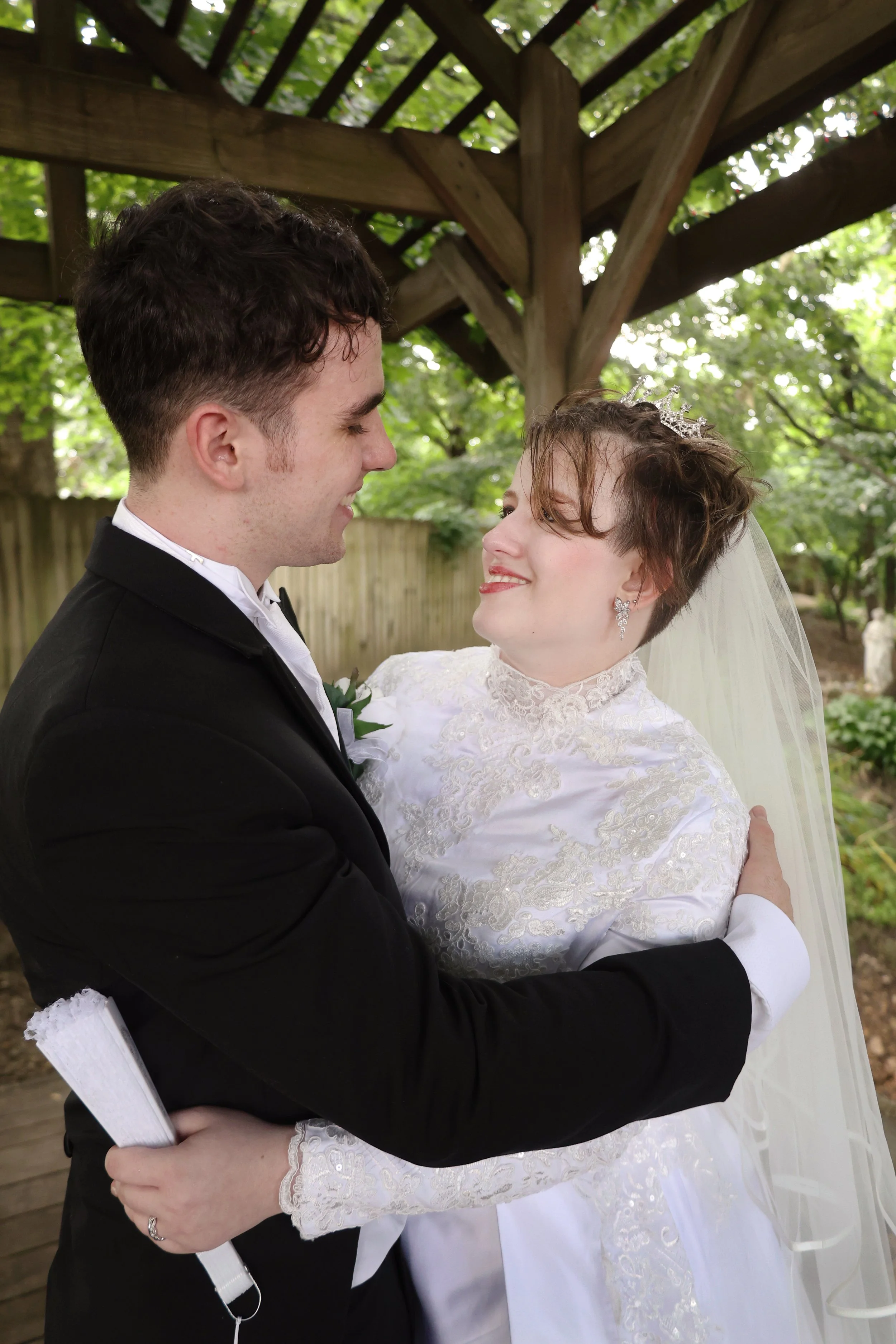A bride and groom smiling and looking at each other, outdoors under a wooden pergola with green leaves in the background, dressed in wedding attire.
