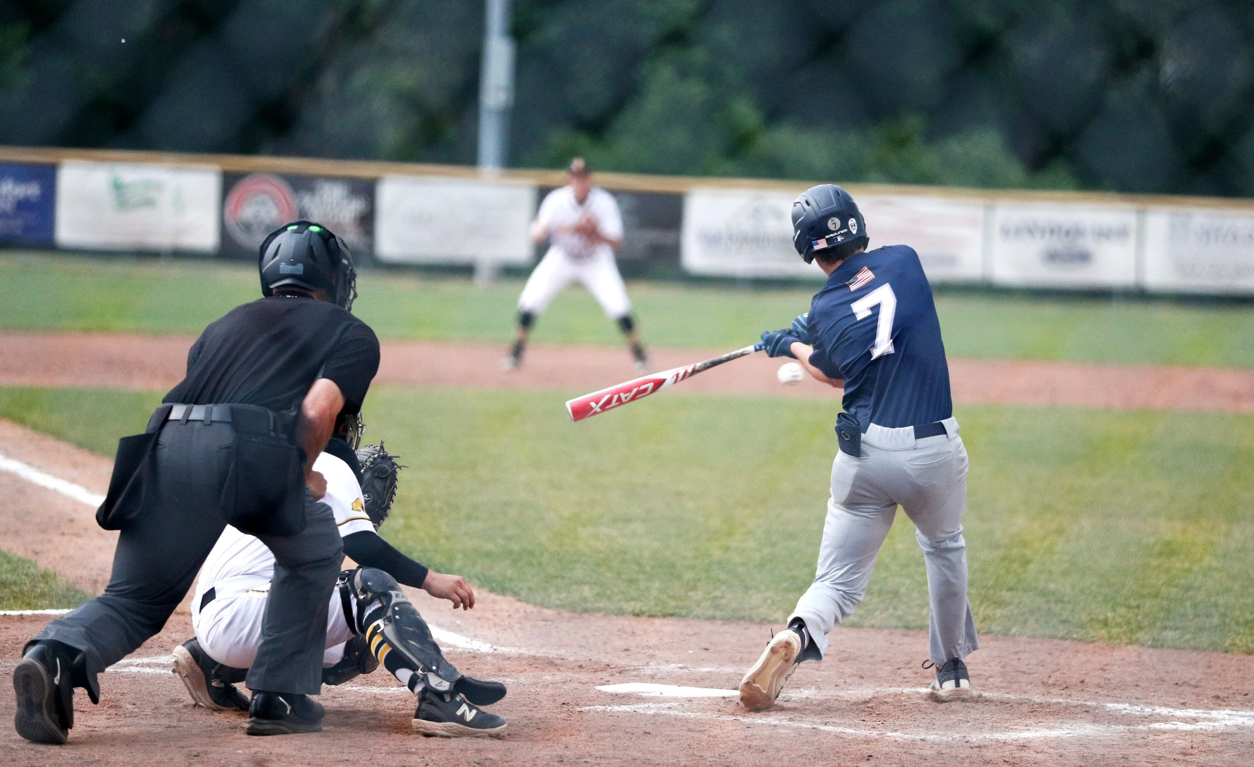 A baseball game in progress with the batter hitting the ball, the catcher and umpire behind home plate, and the pitcher in the background.