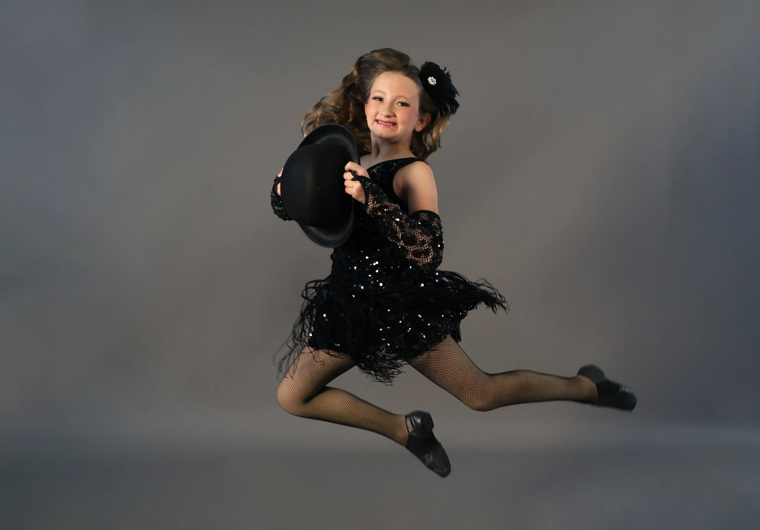 Young girl in a black, sequined dance costume with fishnet tights, holding a black top hat, posing mid-air against a plain gray background.