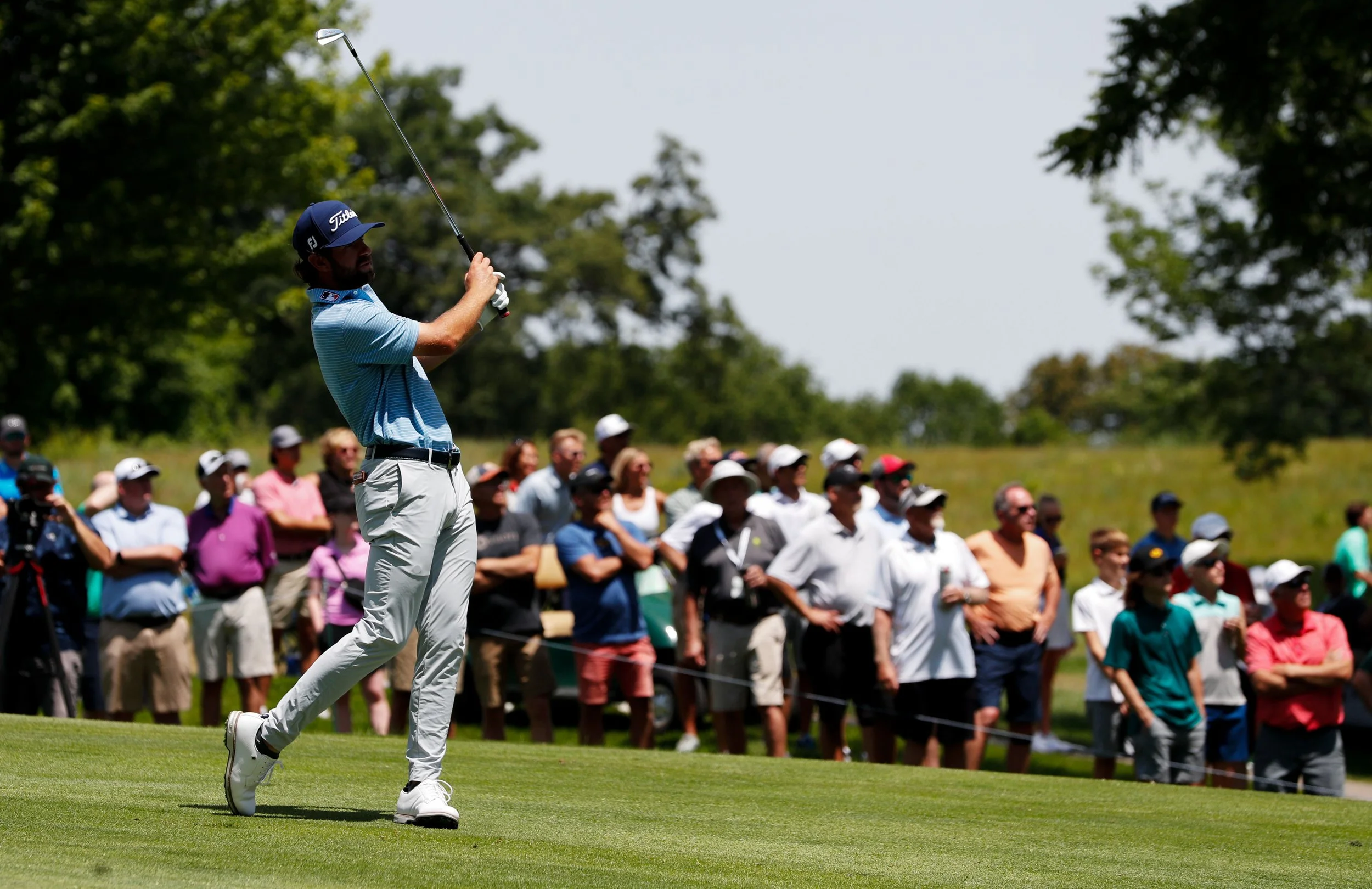 A man in a light blue golf shirt, khaki pants, and a dark blue Titleist cap swings a golf club on a lush green golf course during daytime. In the background, a large crowd of spectators, many wearing hats and sunglasses, watches the game amid trees a