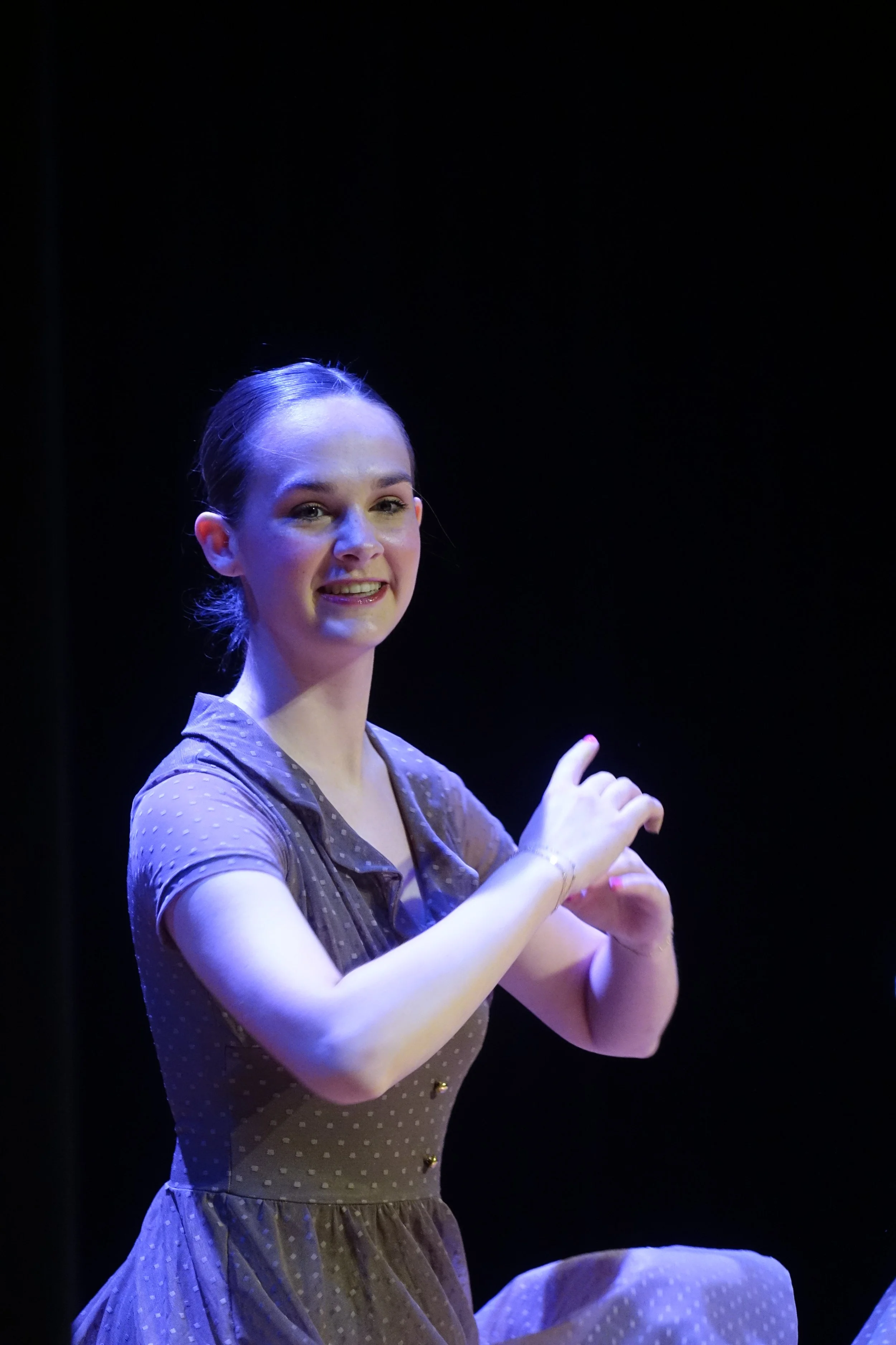 A young woman with light skin and brown hair pulled back, smiling and pointing with her right hand, wearing a brown polka-dot dress on a dark stage with blue lighting.