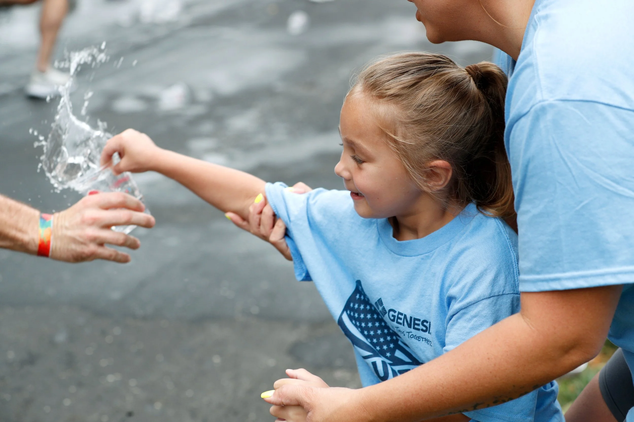 A young girl with a ponytail, wearing a light blue T-shirt and yellow nail polish, smiles as she participates in an outdoor water activity. An adult helps her hold a plastic bag filled with water near a body of water, with other people visible in the