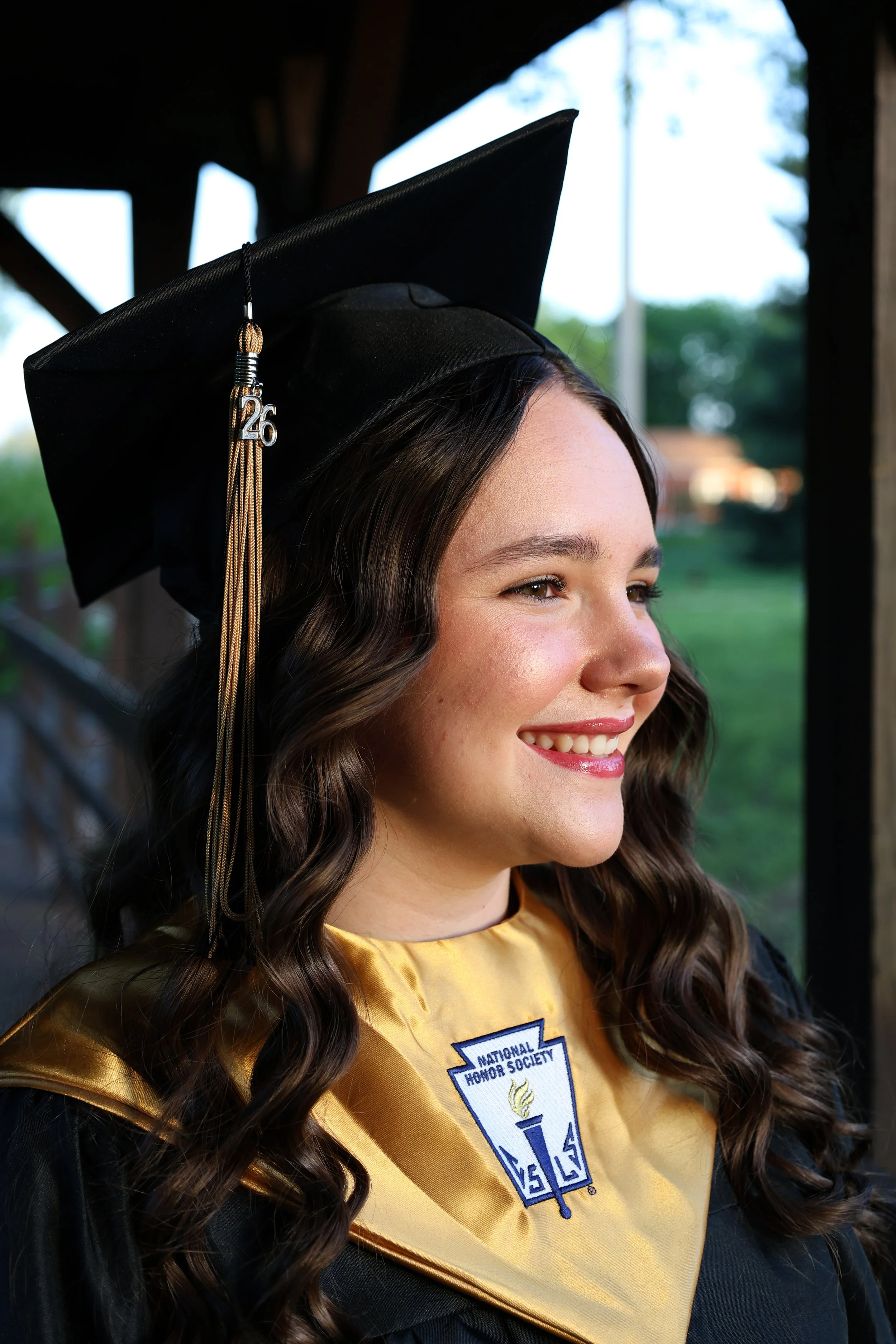 Close-up of a young woman smiling in graduation cap and gown, outdoors with green blurred background.