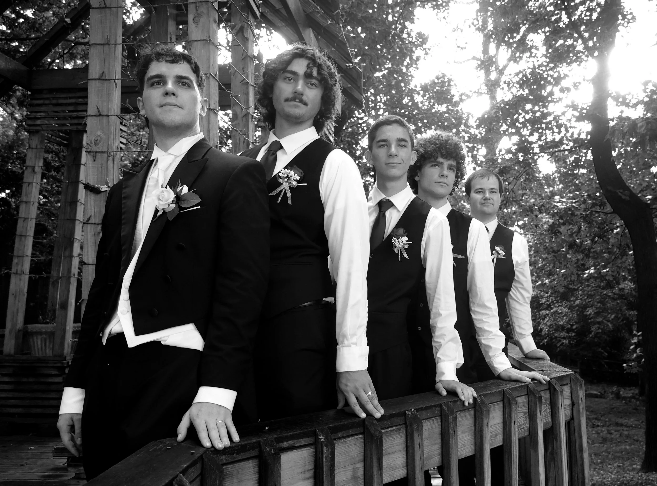 Five young men dressed in formal tuxedos and vests, standing outdoors by a wooden fence with trees and sunlight in the background. The photo is in black and white.