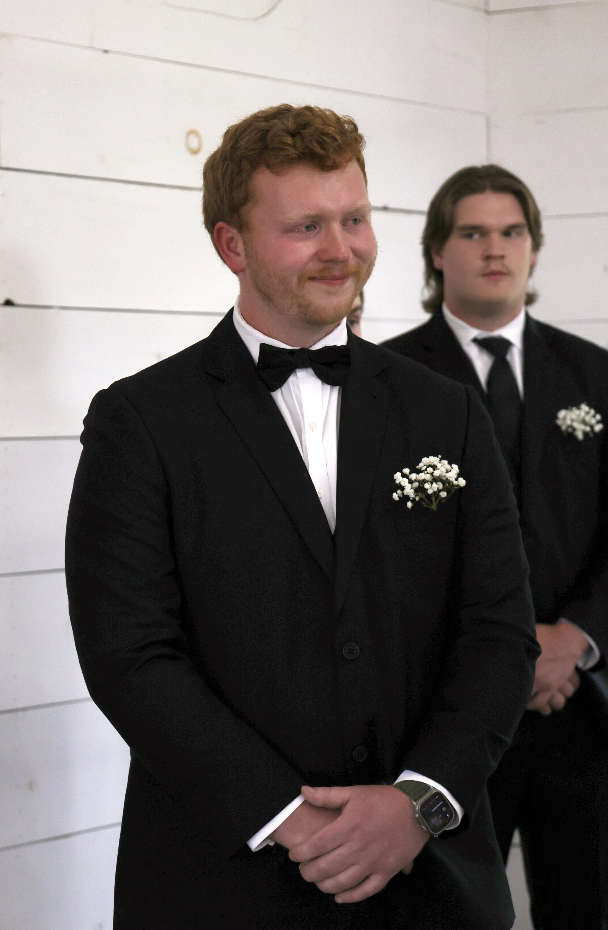 A man in a black tuxedo with a white shirt and black bow tie, wearing a smartwatch, standing with hands clasped, at a formal event. Behind him, another man in formal attire stands against a white wall.