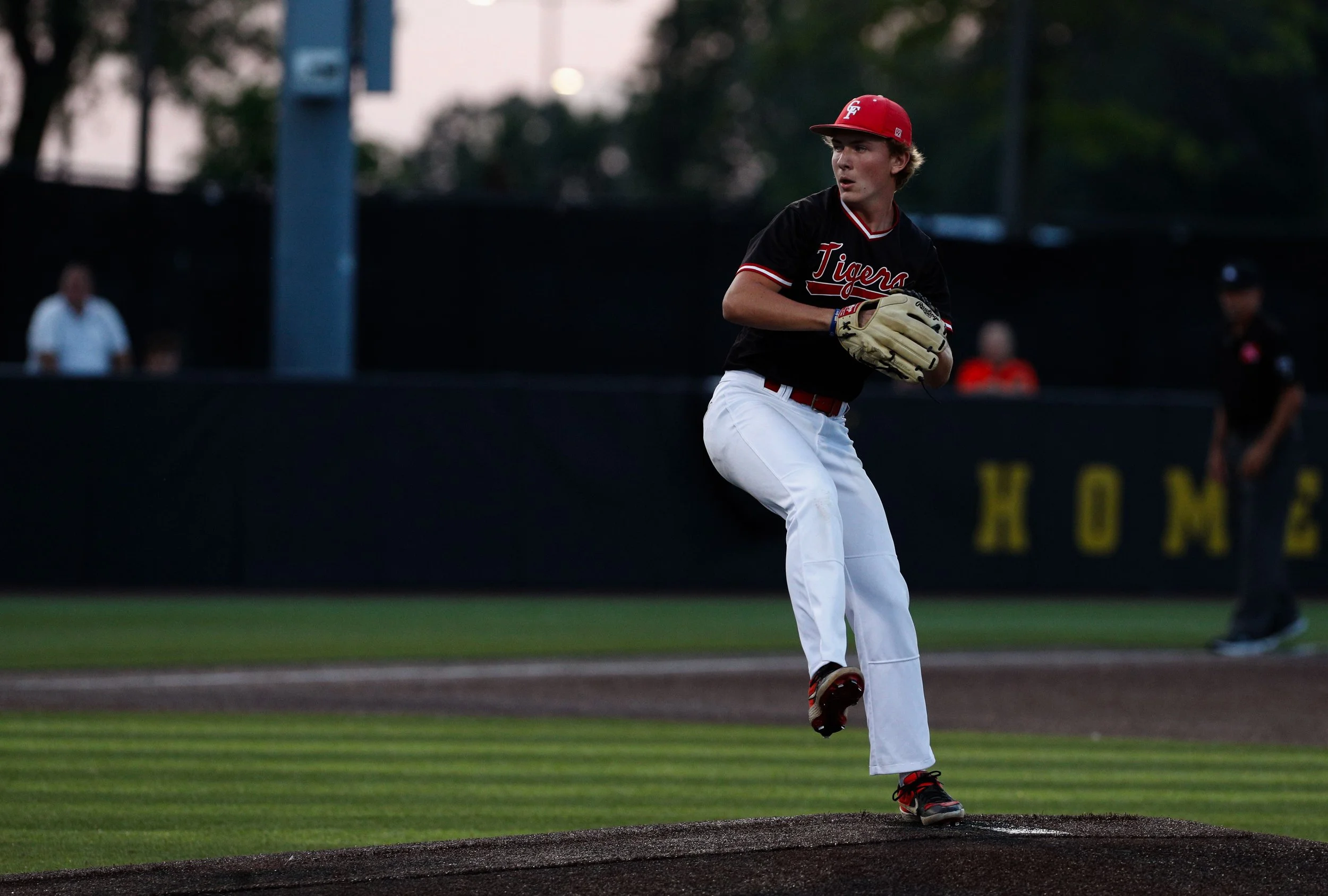 A young baseball player in black and red uniform with a red cap, white pants, and black and red cleats, standing on a baseball mound during a game, holding a baseball glove, with a blurred background of spectators and a stadium.