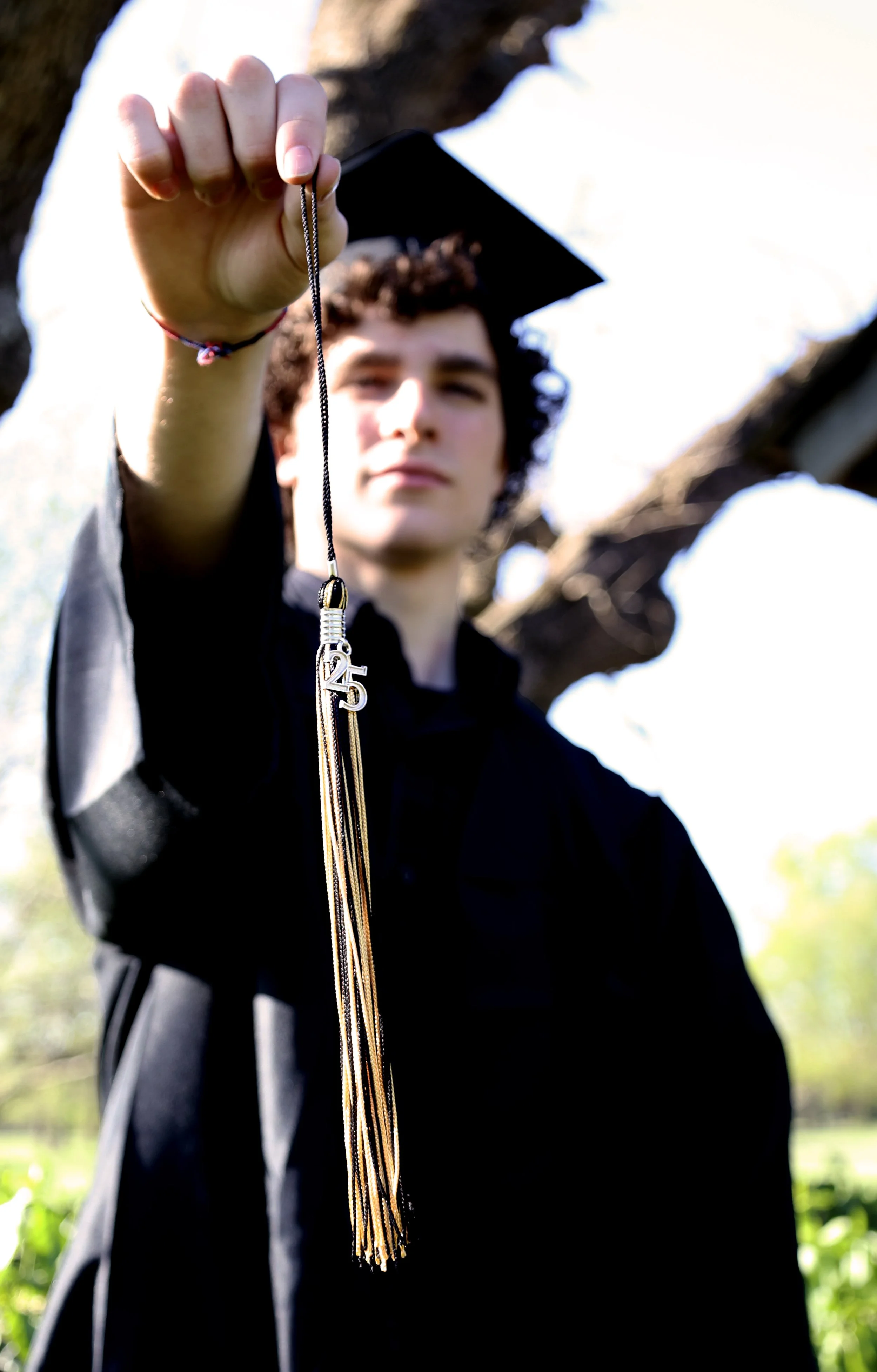 Young person in a graduation cap and gown holding a gold tassel with the number 25, outdoors with trees in the background.