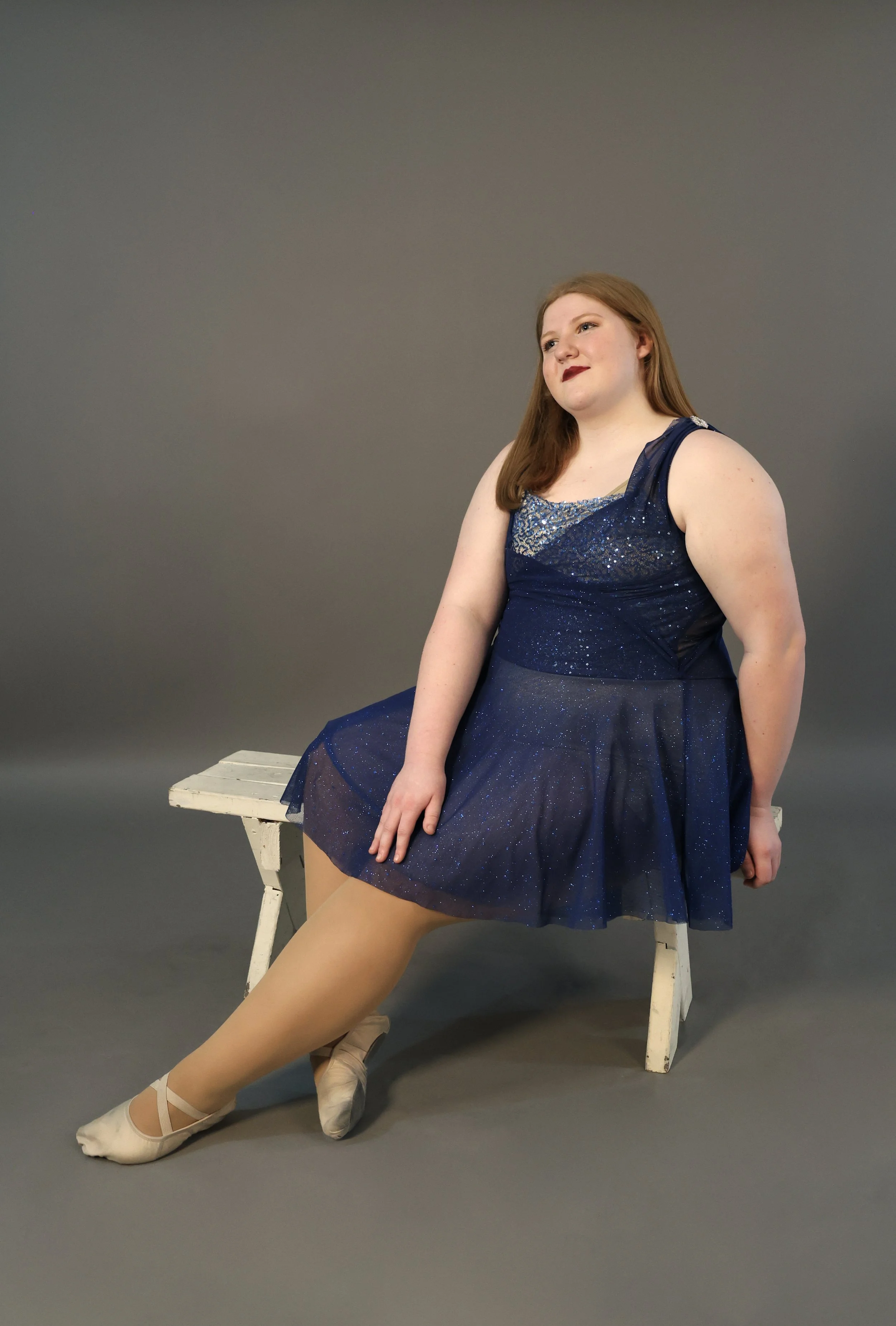 A young woman sitting on a white bench, wearing a blue sparkly ballet dress and pointe shoes, with one leg extended and the other bent, against a plain gray background.