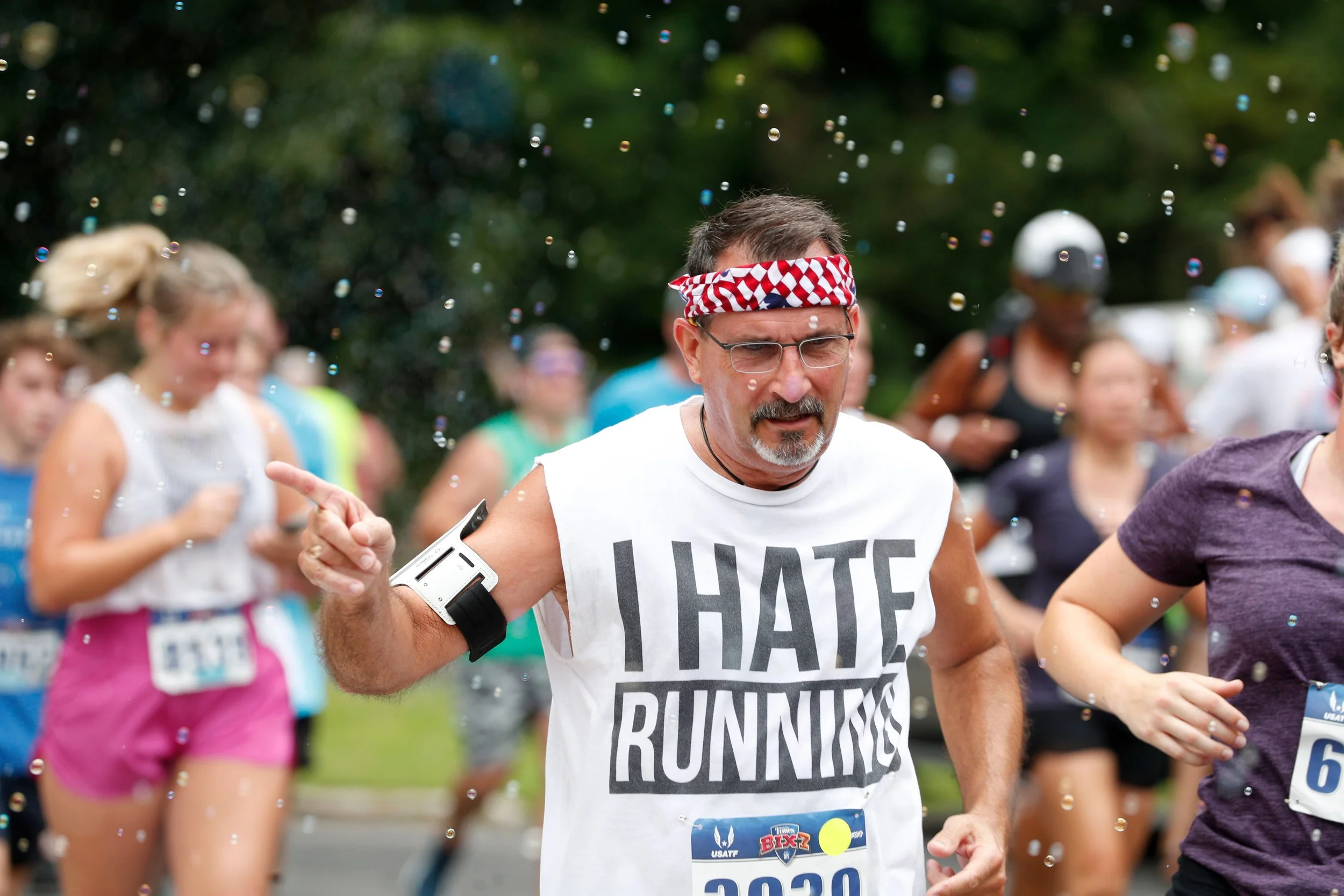 A middle-aged man with glasses, a beard, and a red, white, and blue headband run in a race surrounded by other runners. He wears a white shirt with bold black text and has a serious expression. There are colorful bubbles in the air, and trees in the 