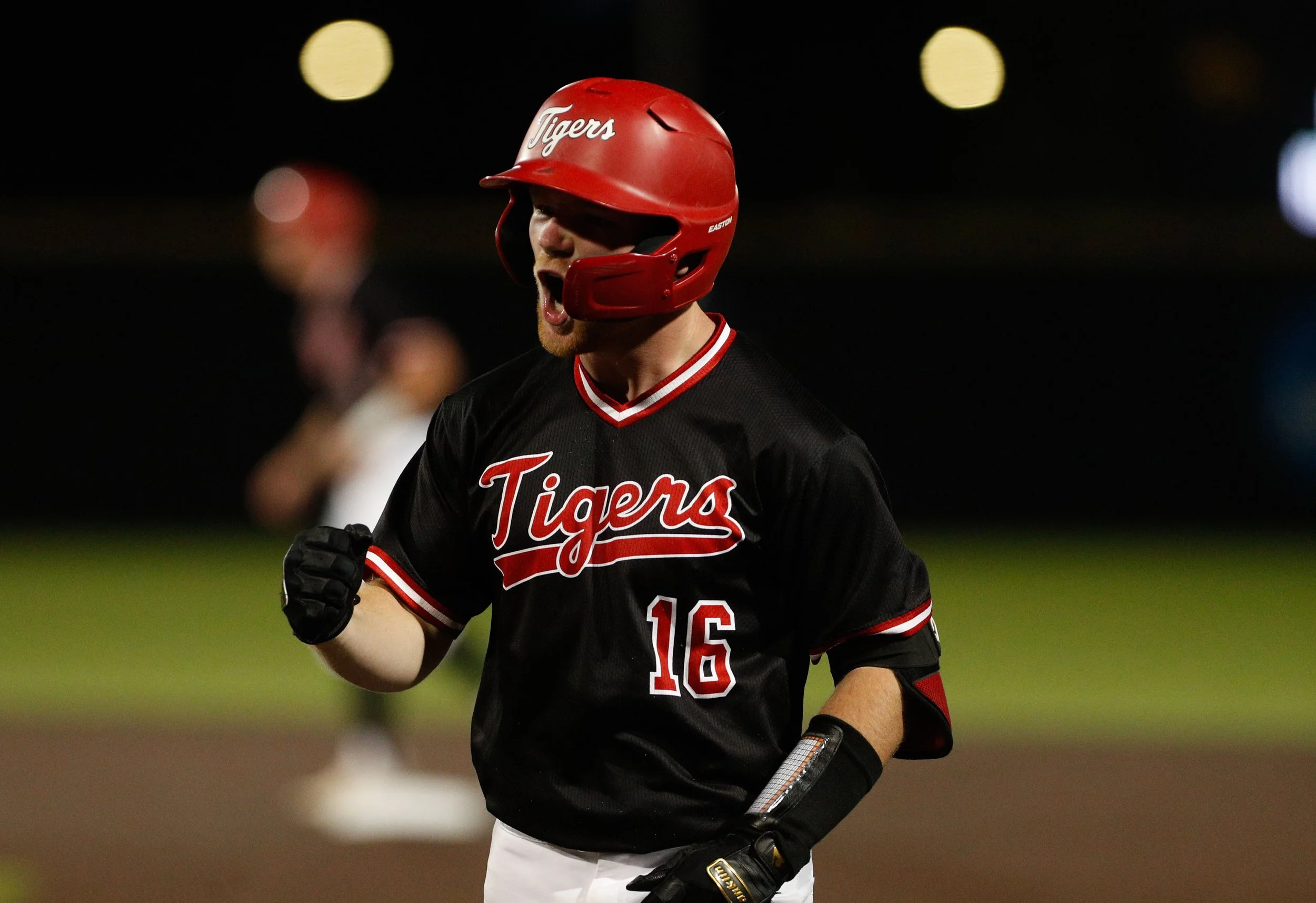 A baseball player in a black and red Tigers uniform with number 16, wearing a red helmet, running or celebrating during a game at night.