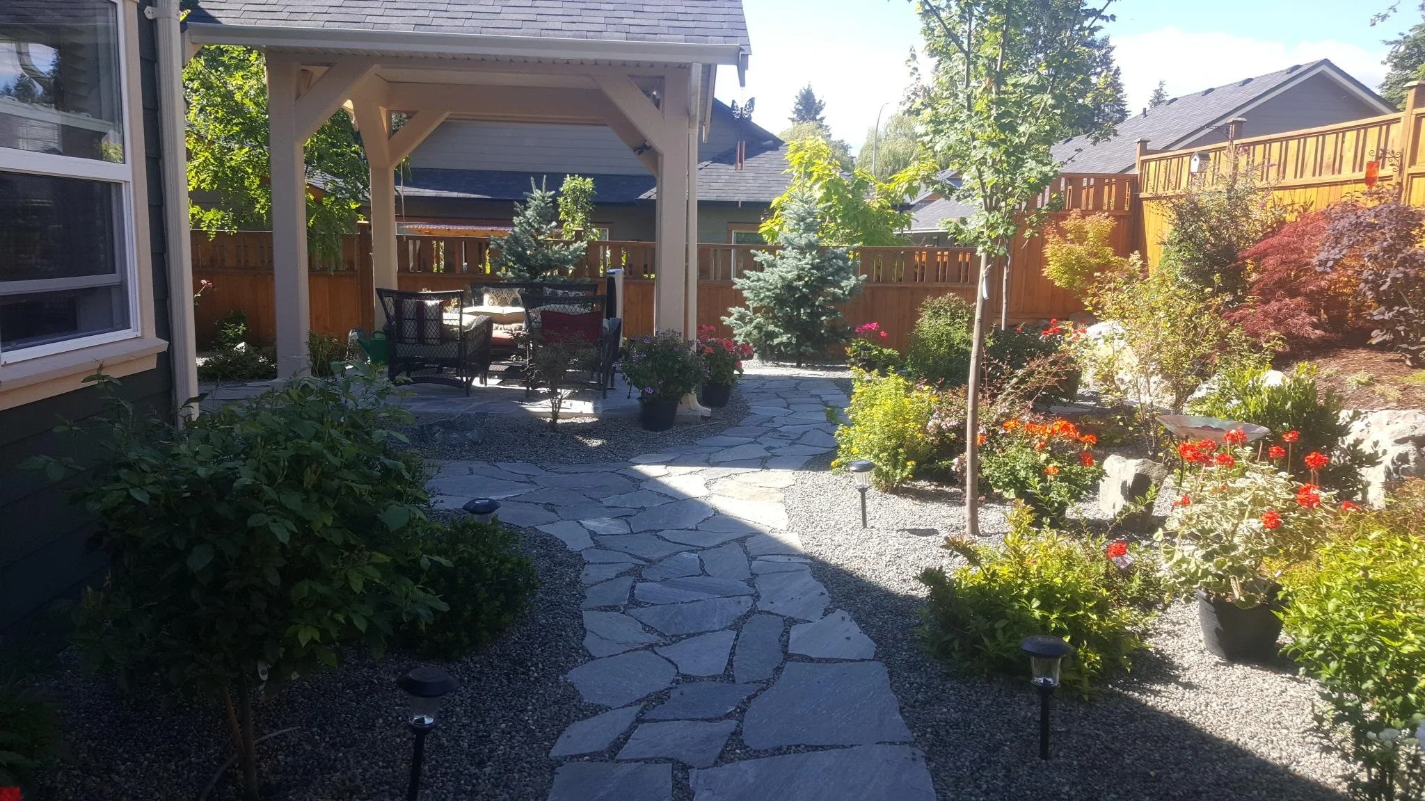 A backyard garden with a stone pathway leading to a covered patio area with outdoor furniture, surrounded by lush green plants, shrubs, and trees, with a wooden fence in the background.