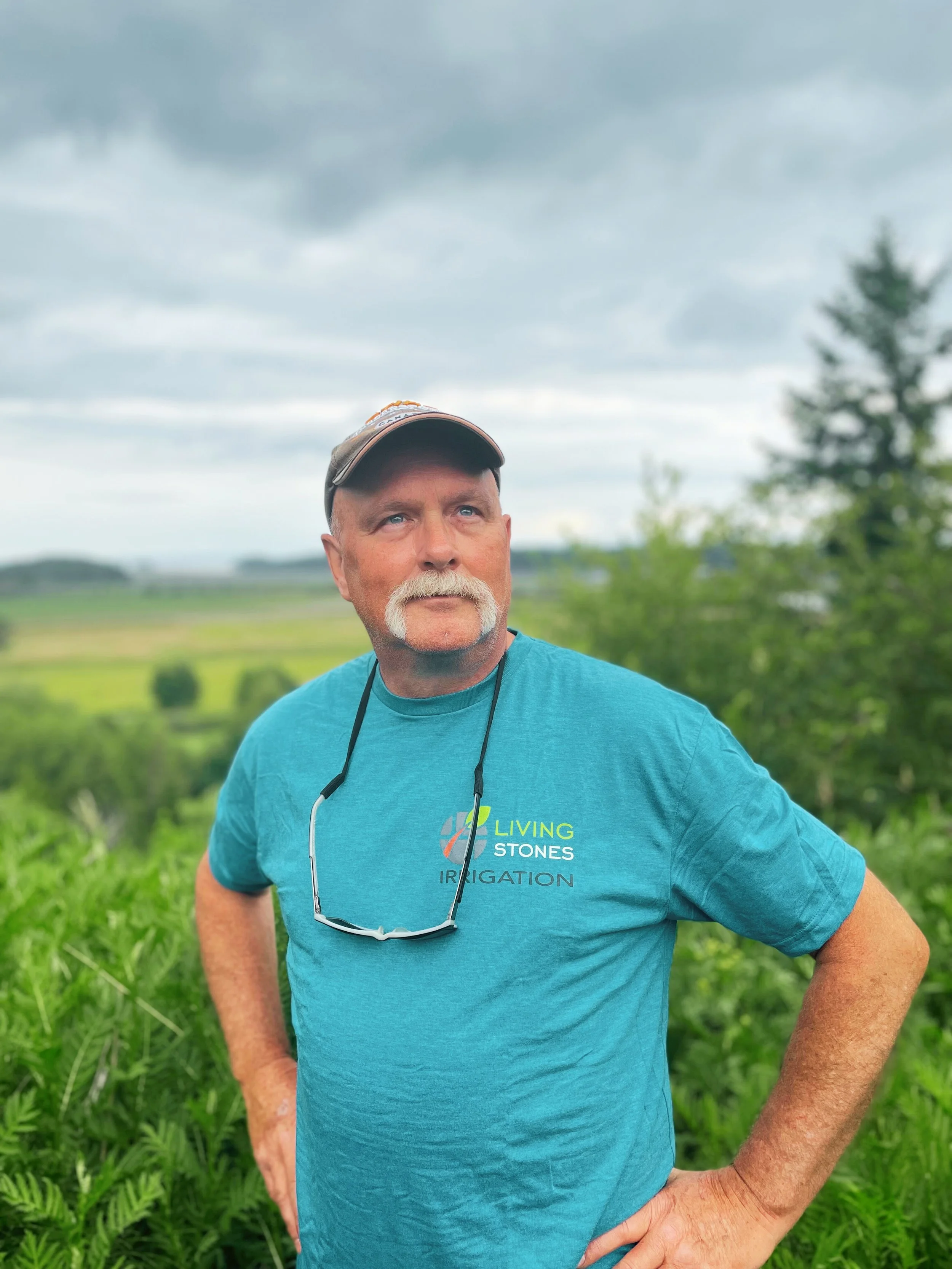 A man with a mustache and beard standing outdoors in a natural setting, wearing a blue t-shirt, a cap, and sunglasses hanging around his neck, with a background of green trees and cloudy sky.
