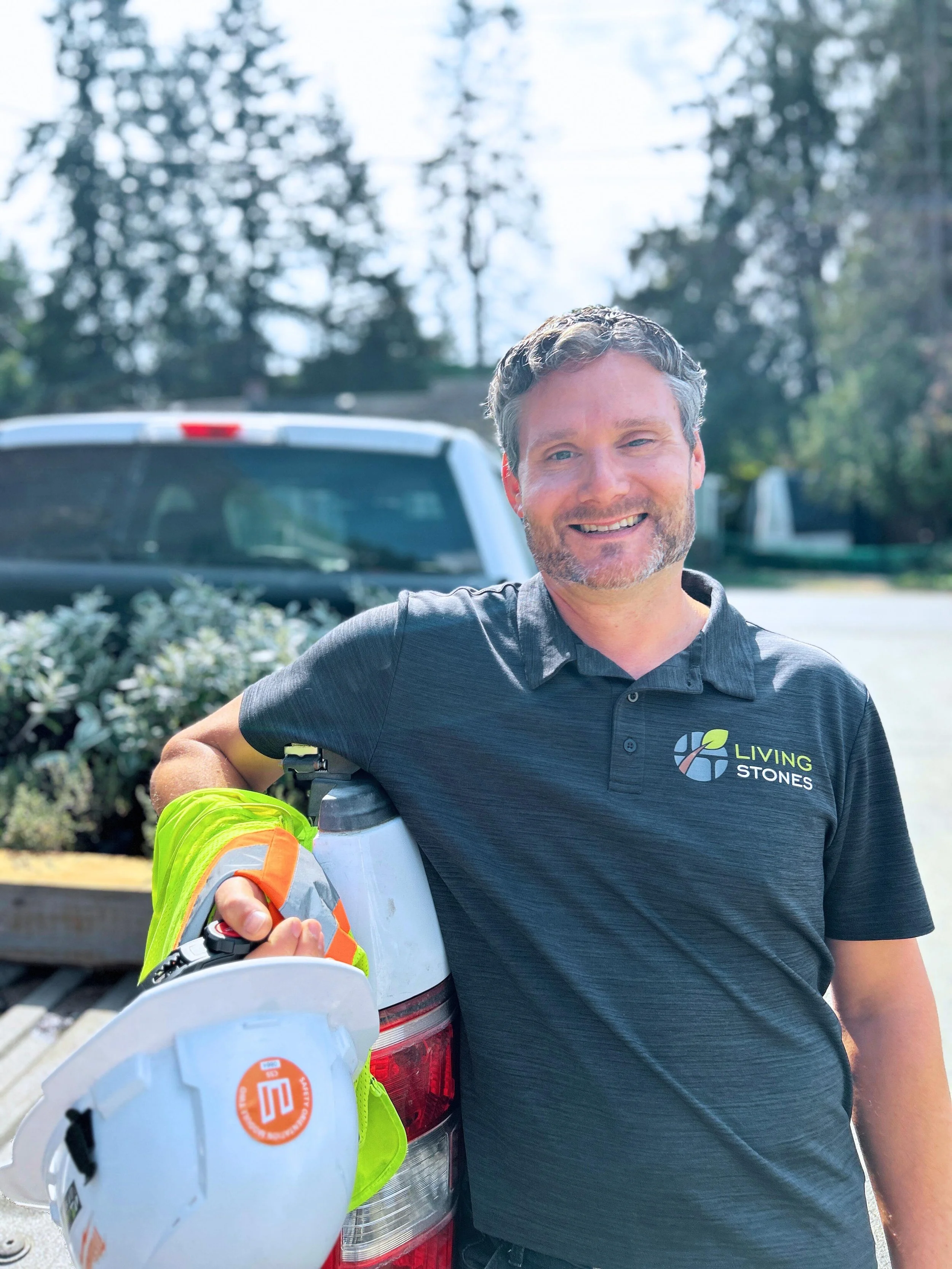 A man leaning on the back of a pickup truck, holding a white hard hat with an orange logo. He is wearing a black polo shirt with a logo that says 'Living Stones' and an illustration of stones and leaves. Background includes trees and a sunny sky.