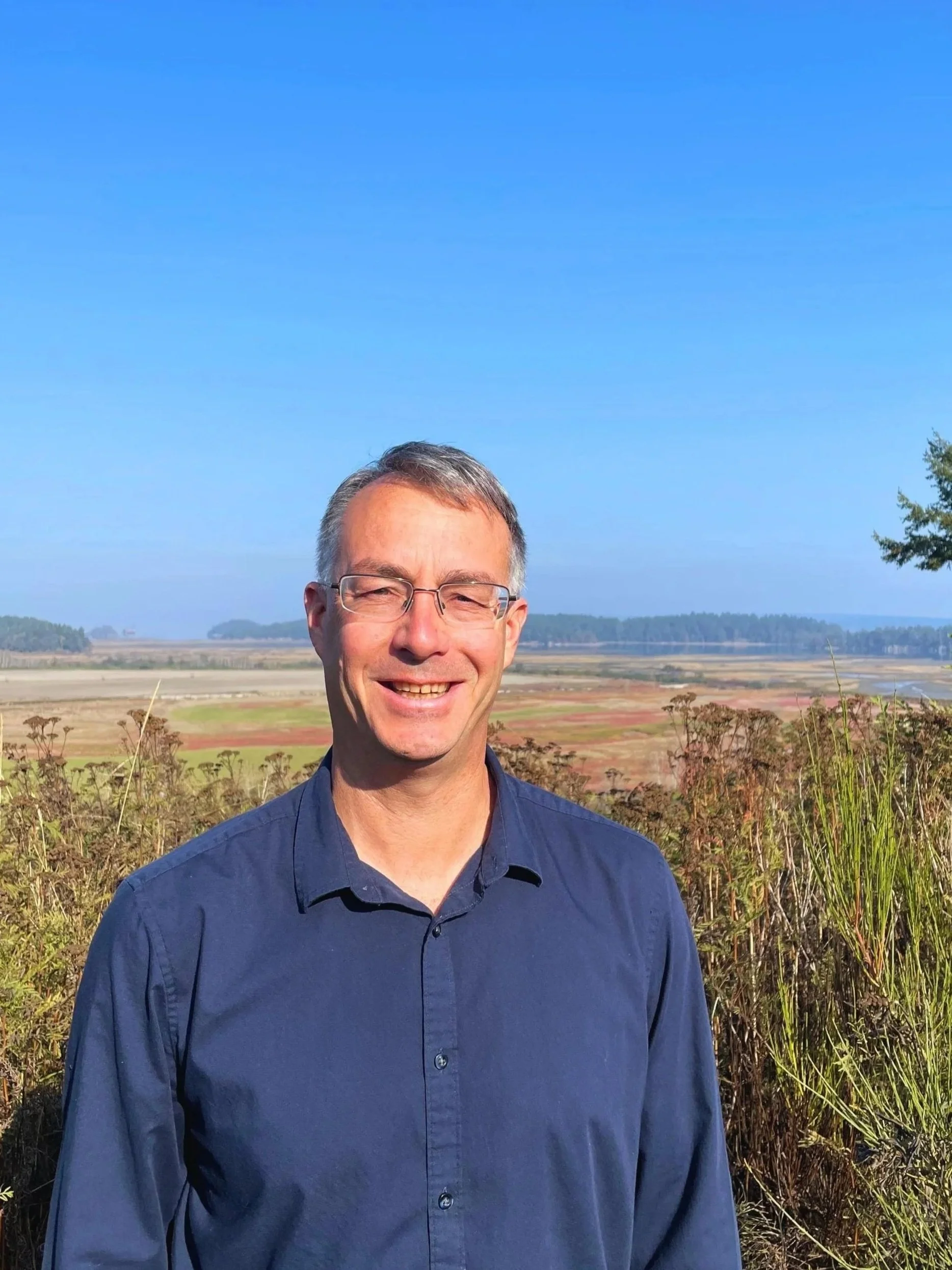 A smiling man in a blue shirt standing outdoors with a rural landscape and clear blue sky in the background.