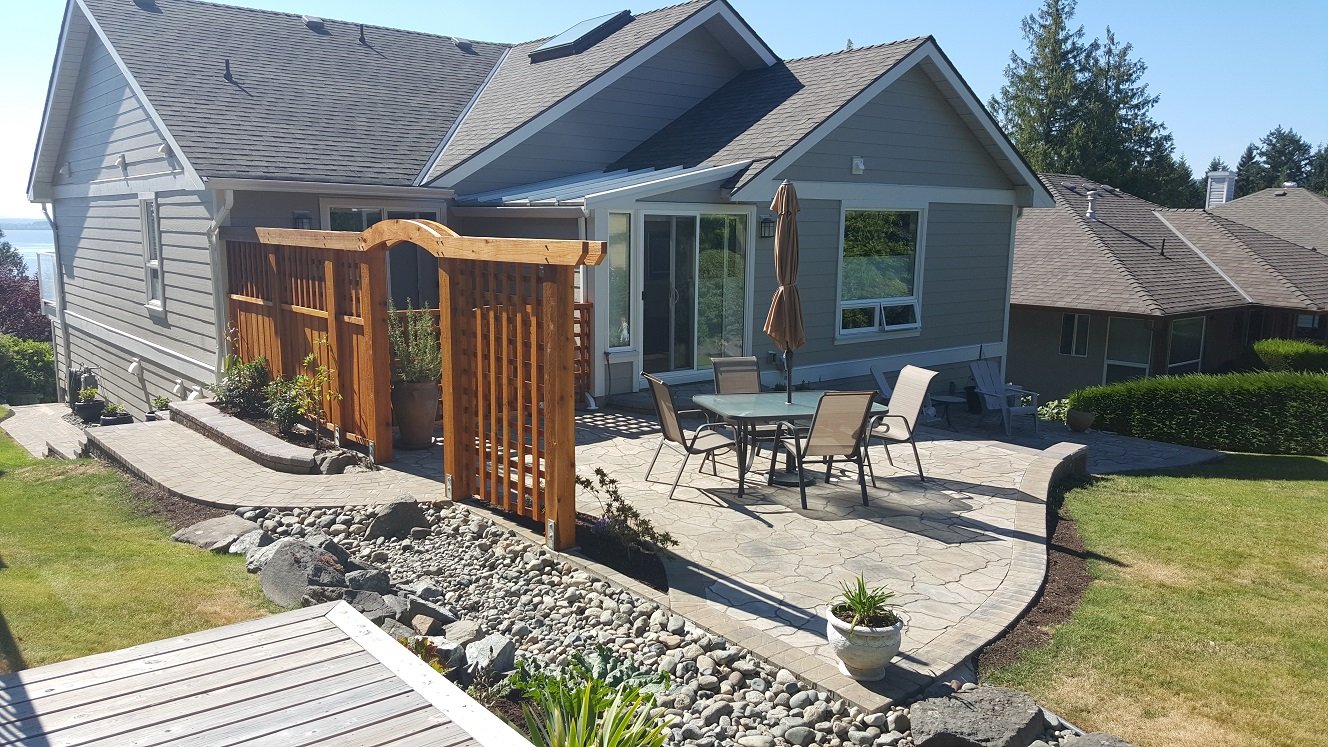 Backyard patio with a wooden fence, outdoor dining table with six chairs, umbrella, and house with sliding glass door and windows.