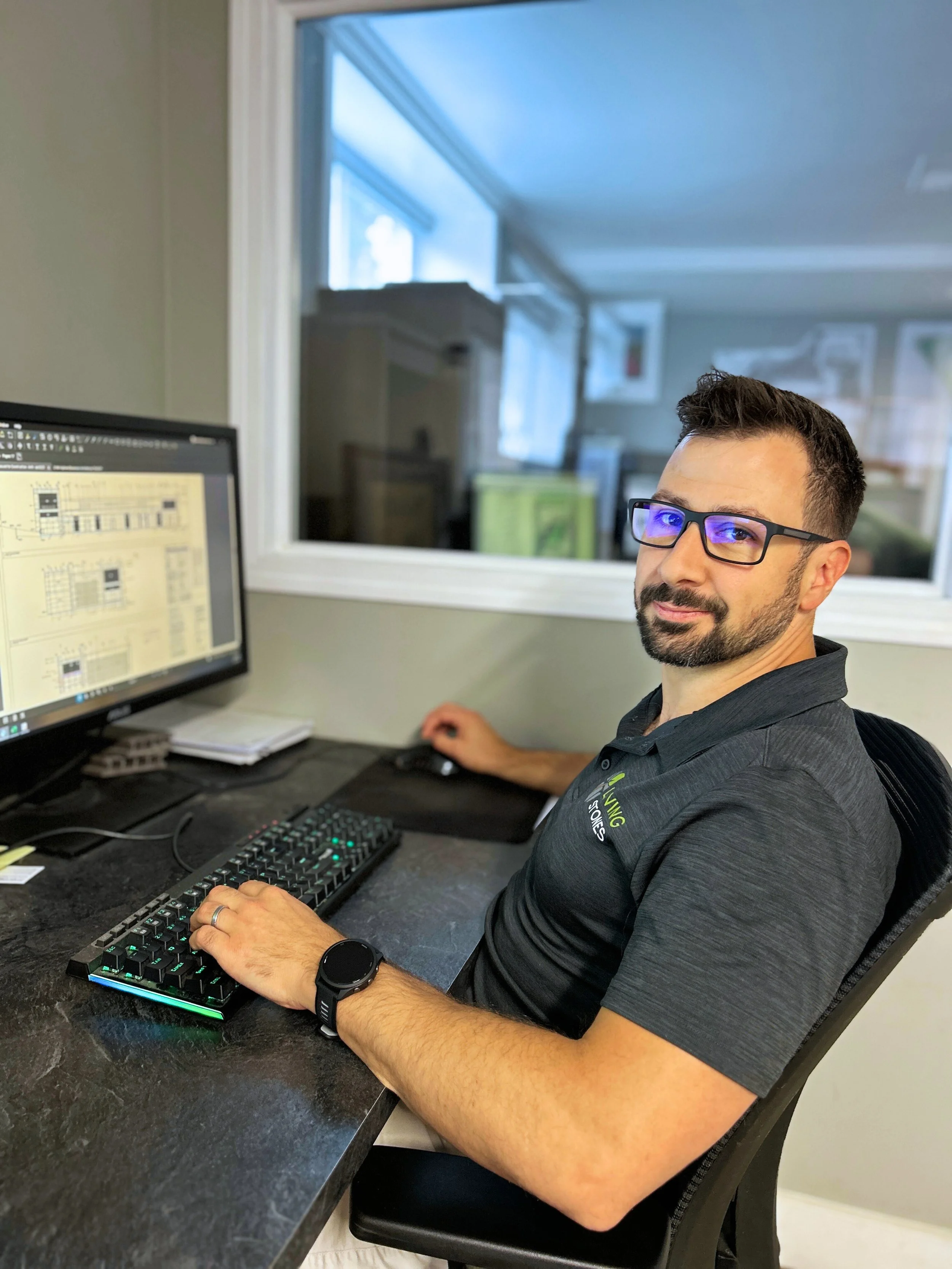 A man with glasses and a beard working at a computer with dual monitors in an office.