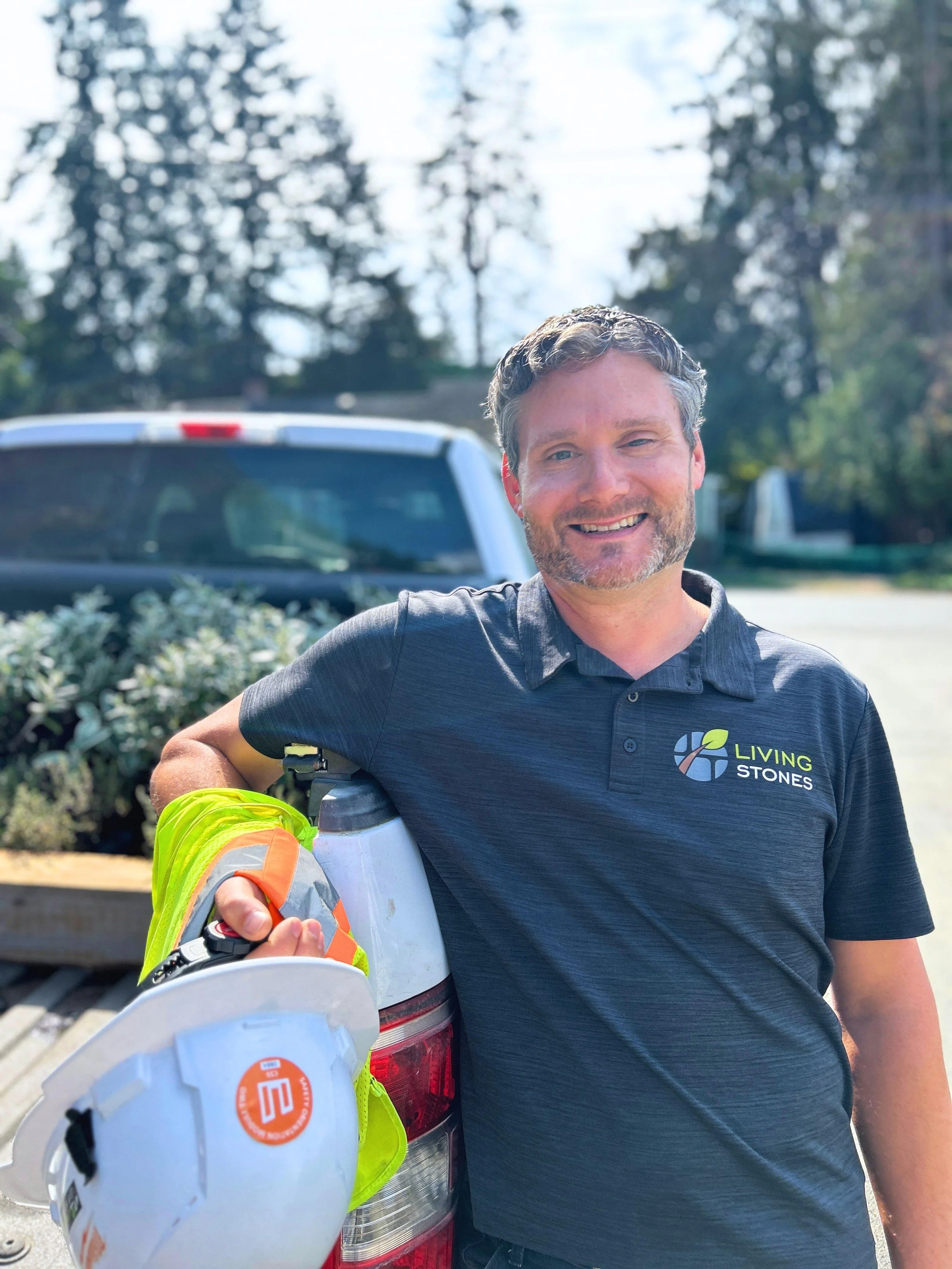 A man leaning on the back of a pickup truck, holding a white hard hat with an orange logo. He is wearing a black polo shirt with a logo that says 'Living Stones' and an illustration of stones and leaves. Background includes trees and a sunny sky.