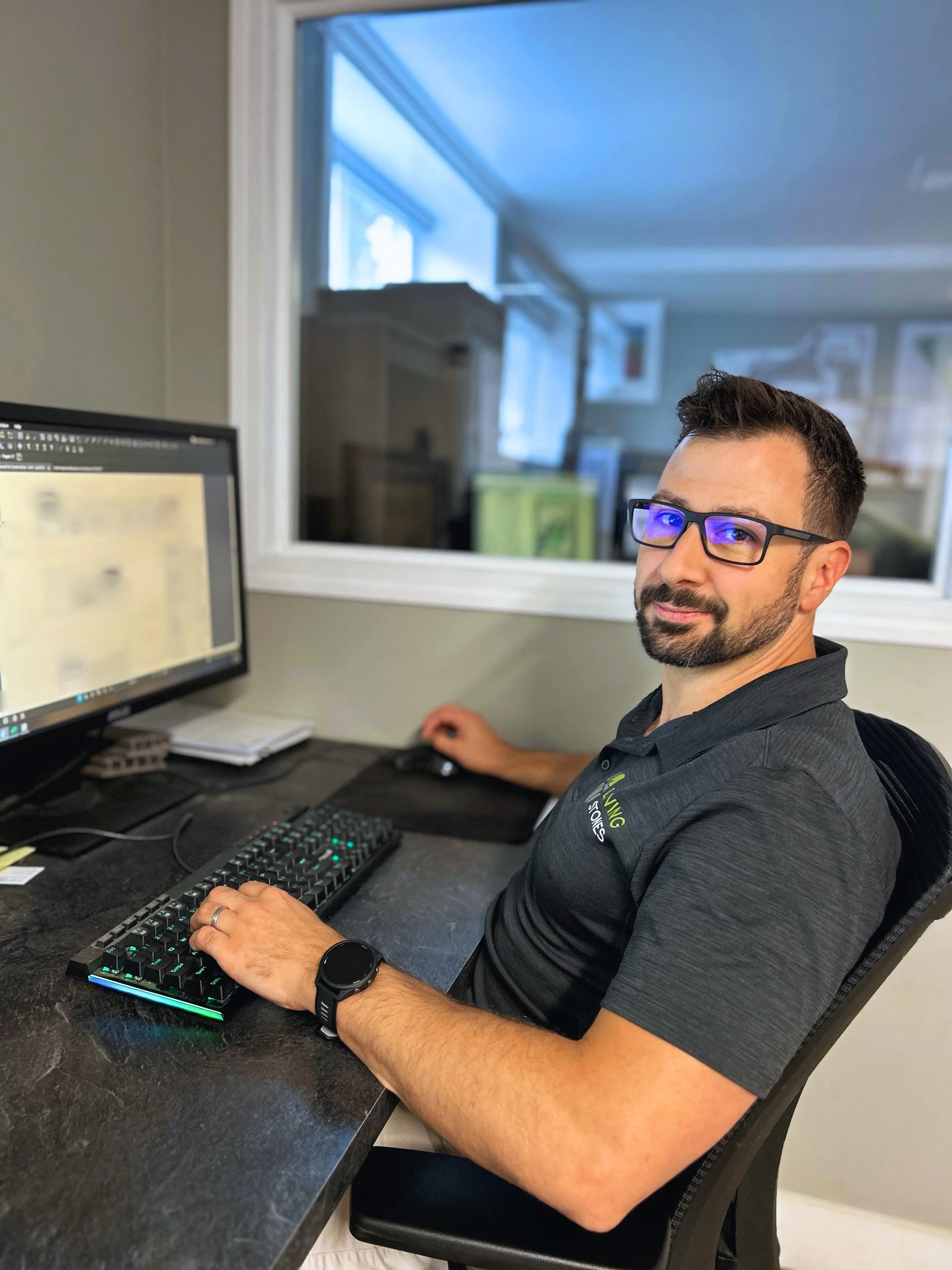 A man with glasses and a beard working at a computer with dual monitors in an office.