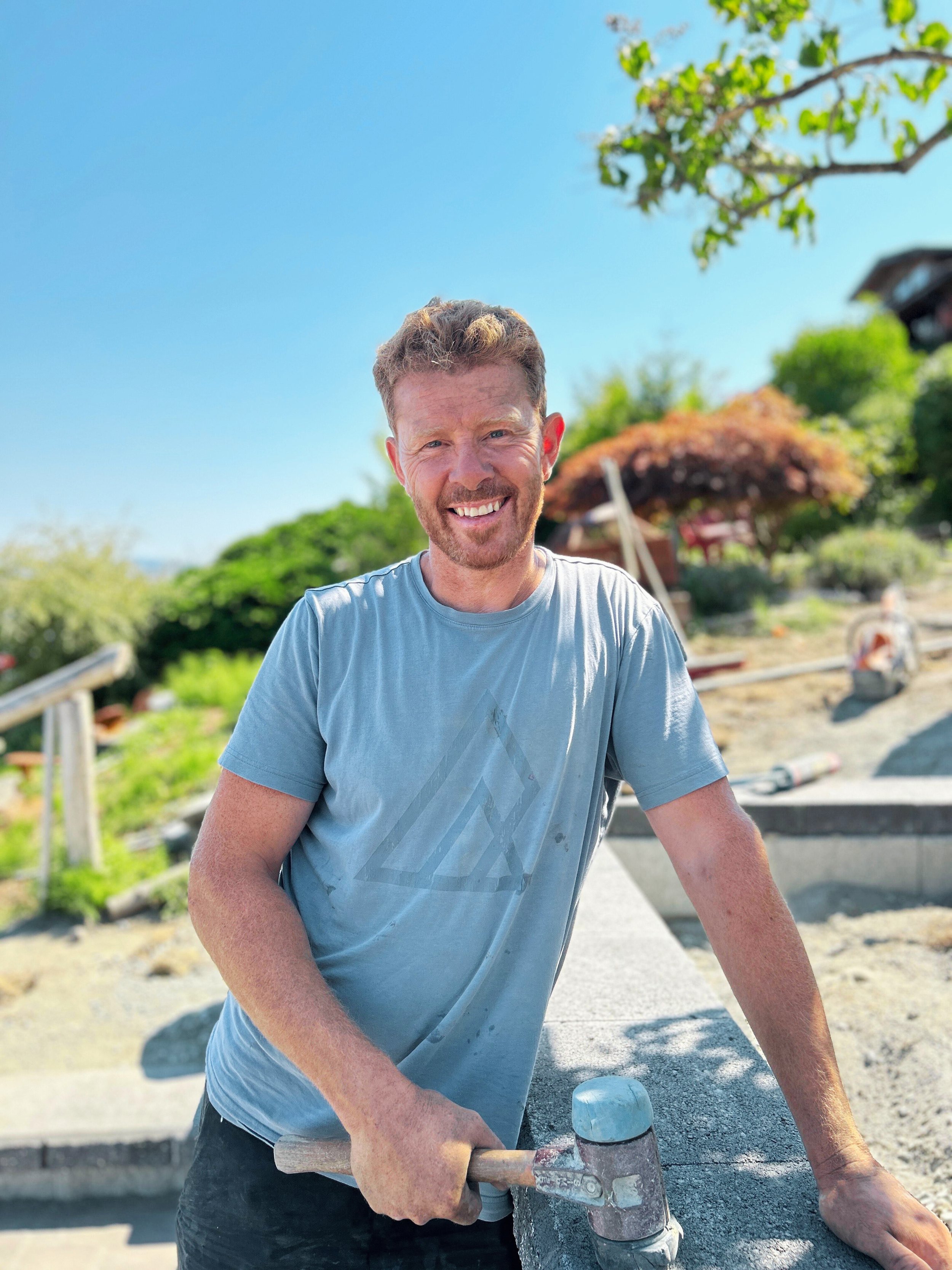 A man smiling at a construction site on a sunny day, holding a hammer with concrete work around him and trees in the background.
