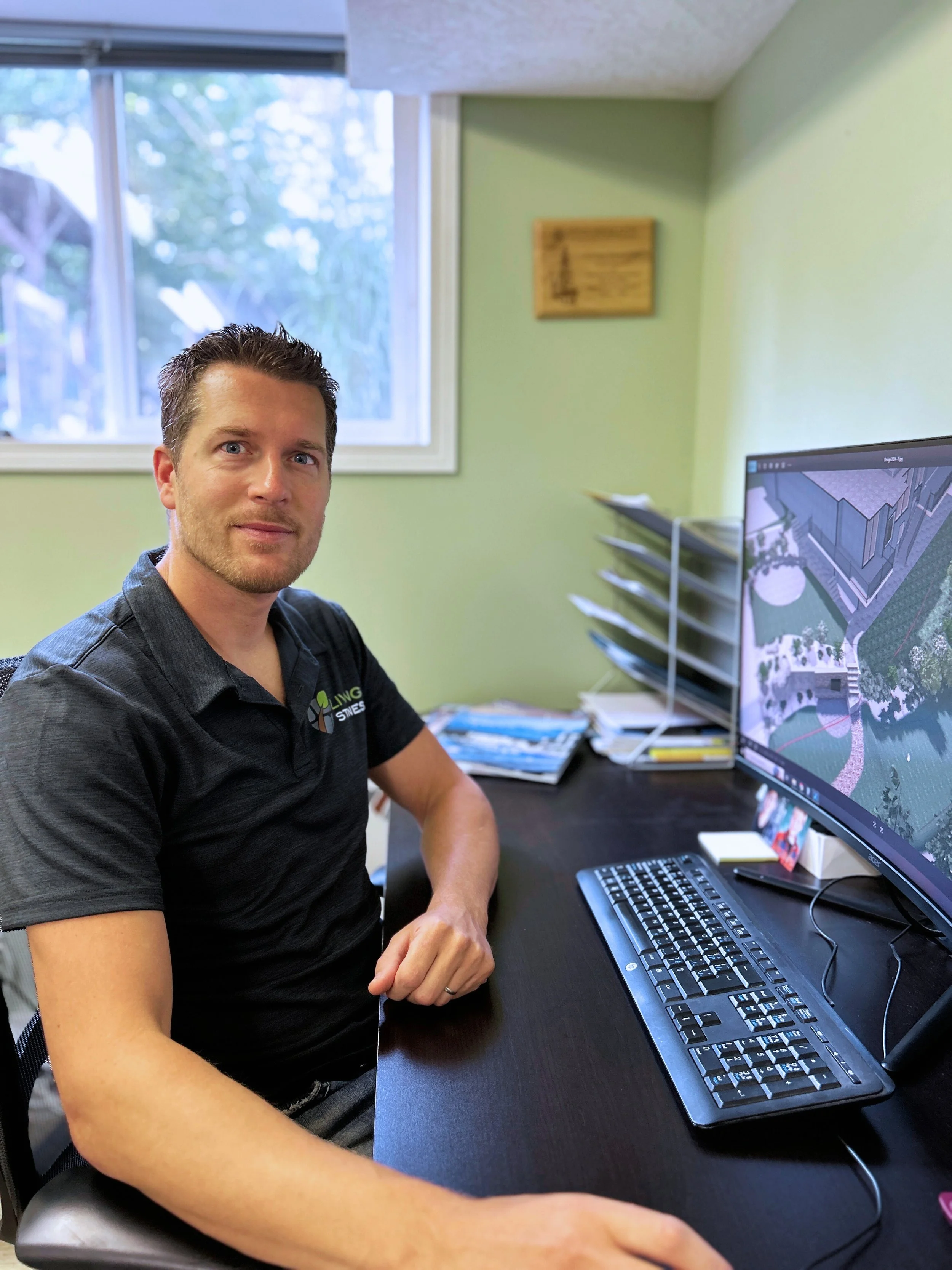 Man sitting at a desk with a computer monitor displaying a landscape design, with office supplies and stacks of documents nearby in a room with light green walls and a window showing trees outside.
