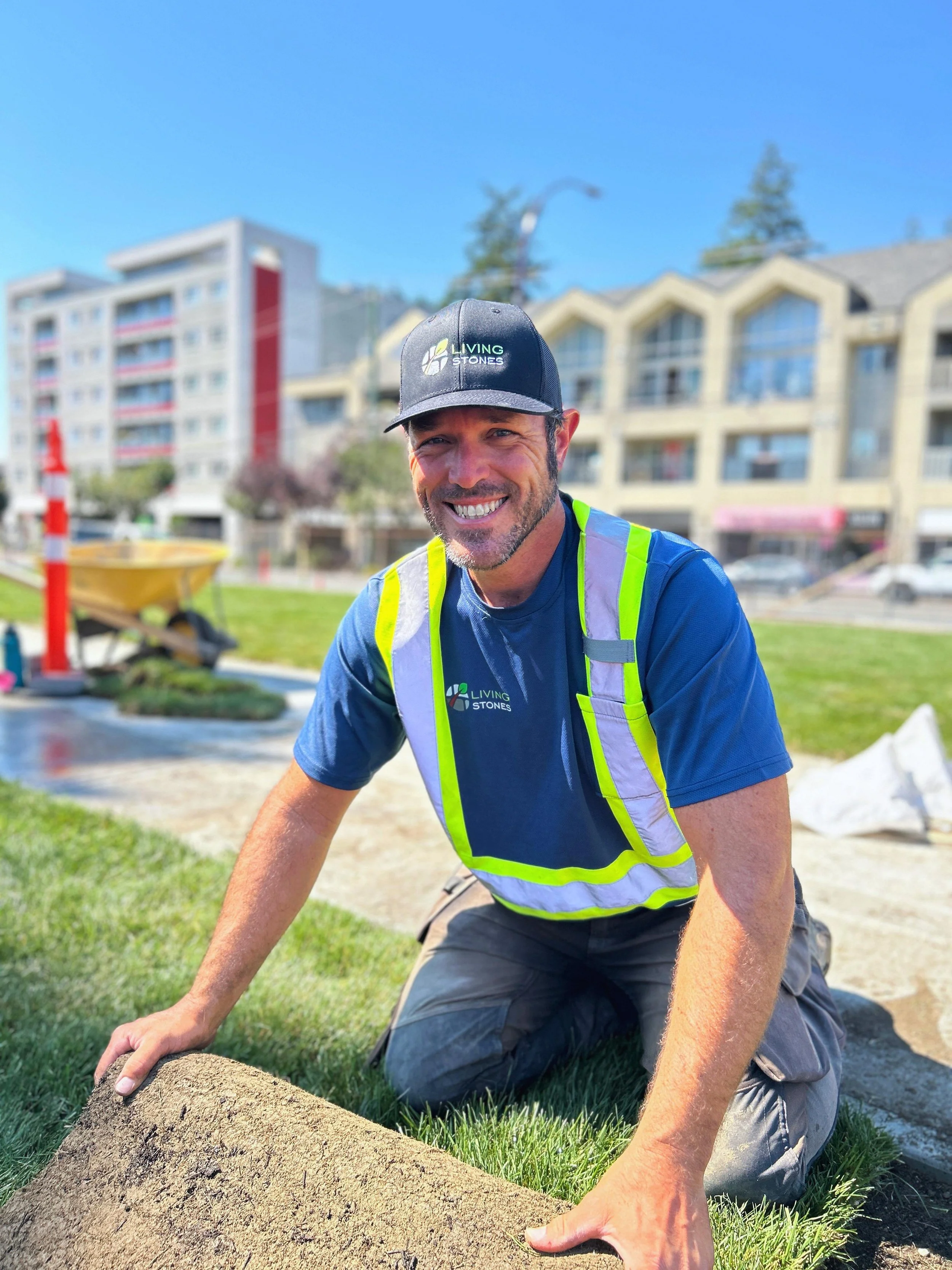 Man kneeling on grass, smiling, wearing a black cap, blue t-shirt with logos, and a reflective safety vest, working on landscaping or construction project outdoors with buildings in the background.