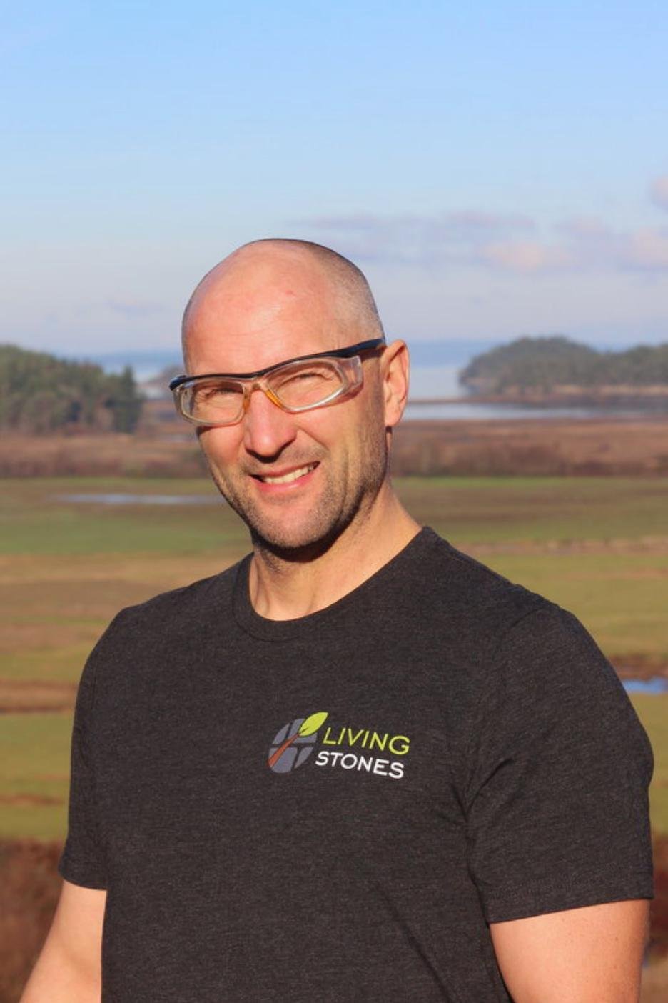 Man with glasses and a beard smiling outdoors with a body of water and trees in the background.