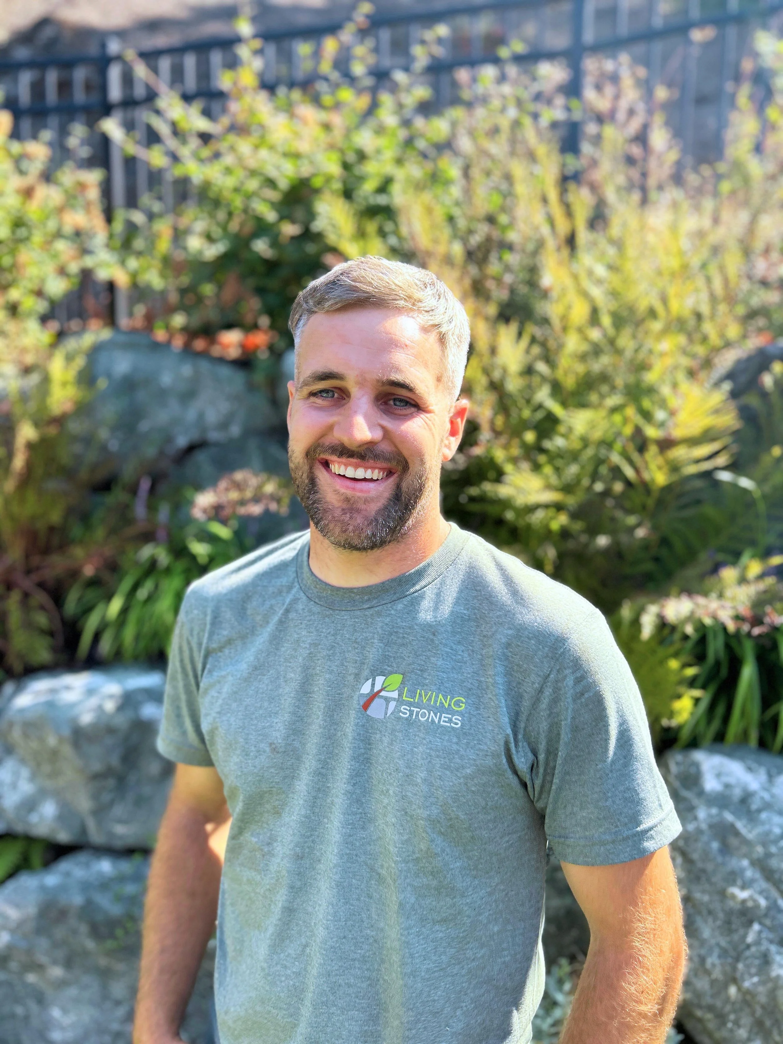 Smiling man with short gray hair and beard, wearing a gray T-shirt with a logo, standing outdoors in a garden with rocks and plants.