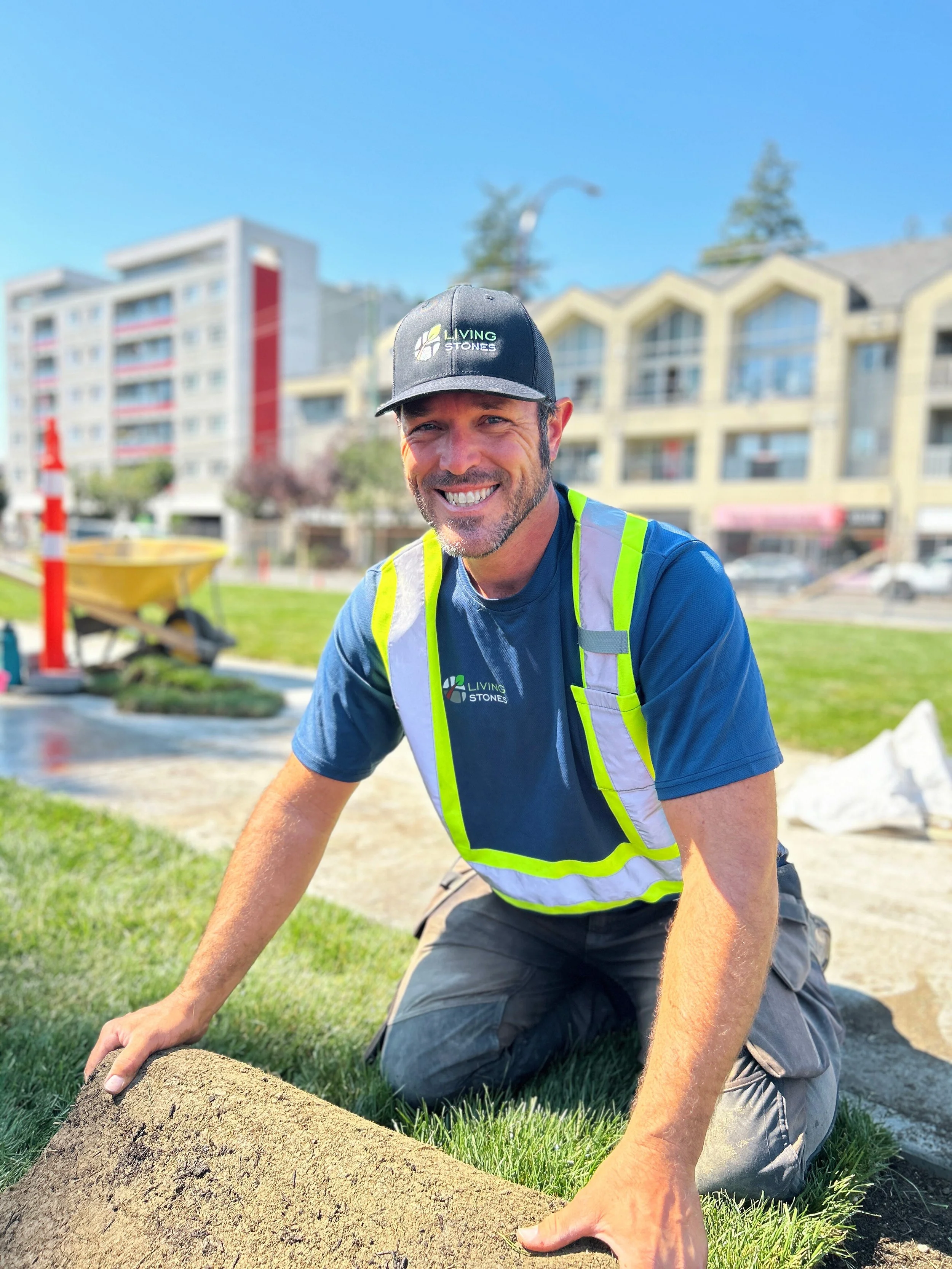 Man kneeling on grass, smiling, wearing a black cap, blue t-shirt with logos, and a reflective safety vest, working on landscaping or construction project outdoors with buildings in the background.