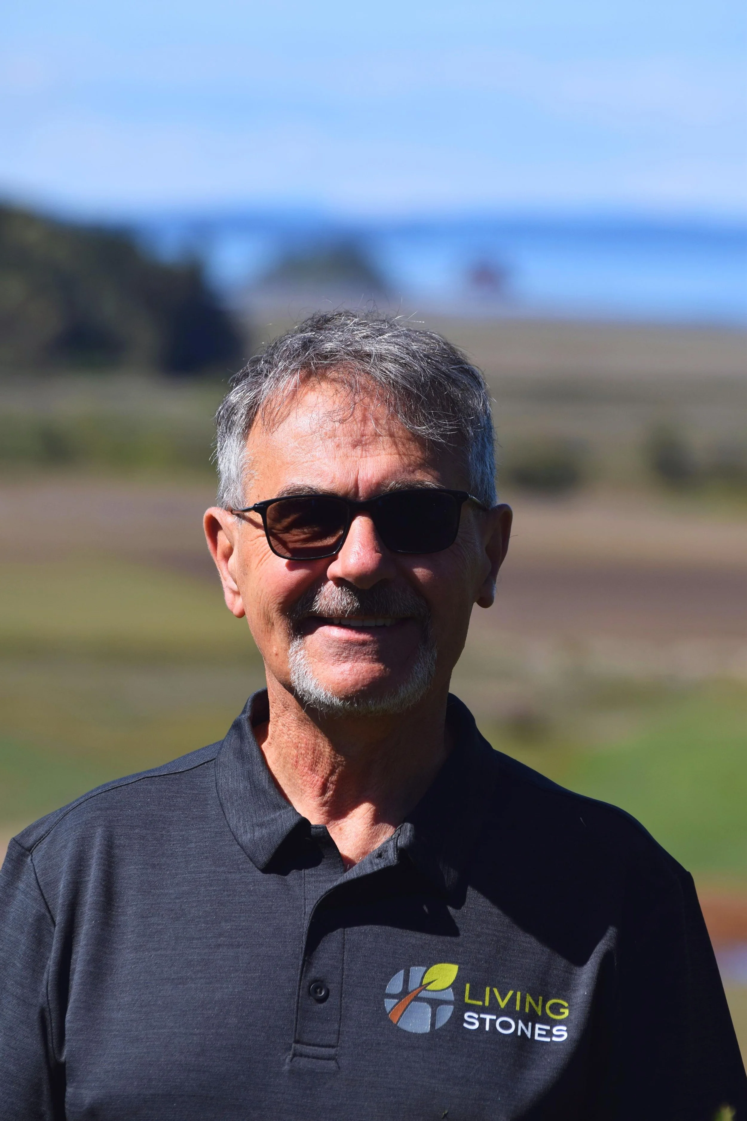 A smiling man wearing sunglasses and a black polo shirt with the 'Living Stones' logo standing outdoors with blurred background of a landscape, possibly a beach or fields.