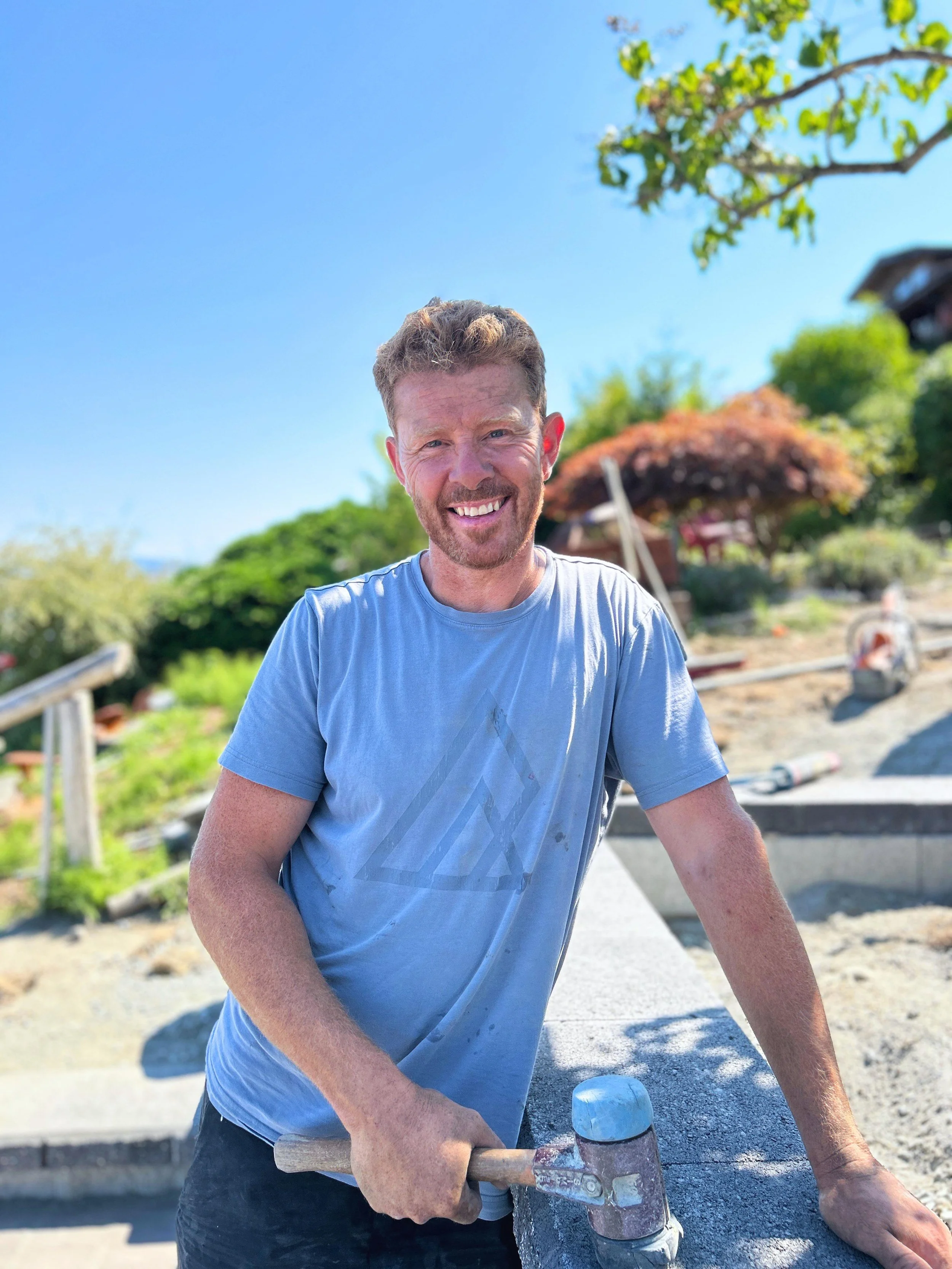 A man smiling at a construction site on a sunny day, holding a hammer with concrete work around him and trees in the background.