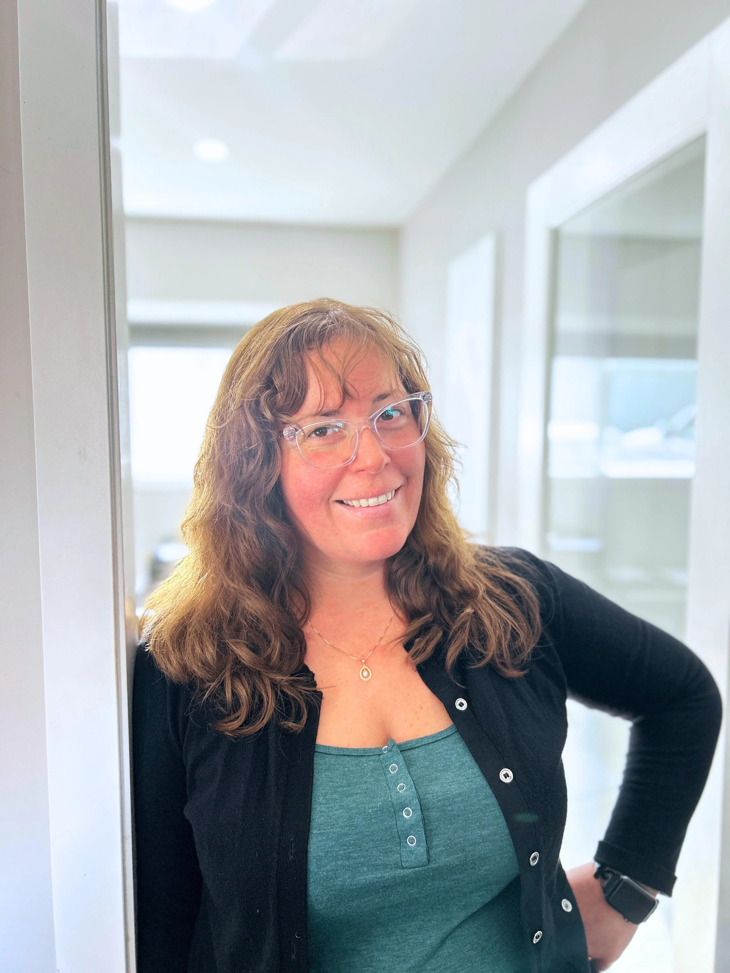 A woman with wavy brown hair, glasses, and a friendly smile standing indoors, wearing a teal top, black cardigan, and a watch on her left wrist.