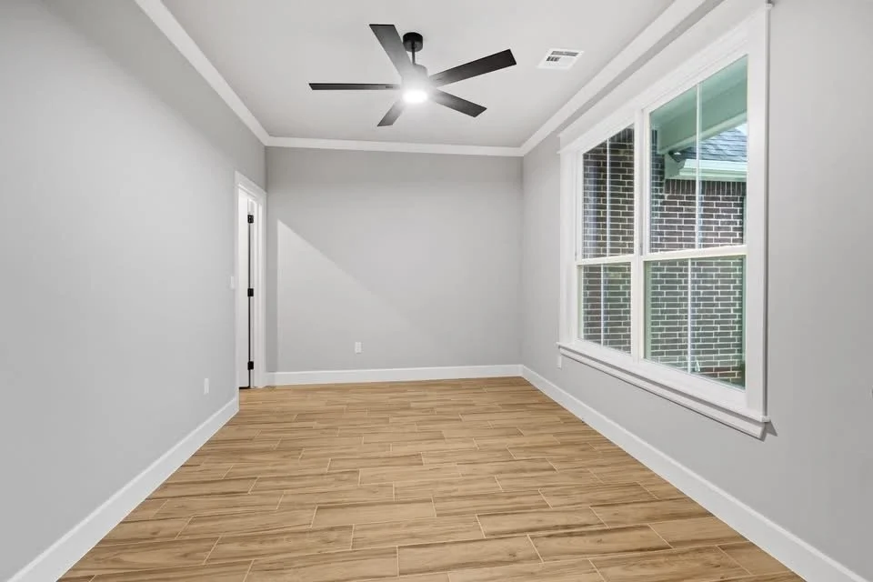 Empty room with light gray walls, white trim, hardwood flooring, a black ceiling fan, and a large window showing brick outside.