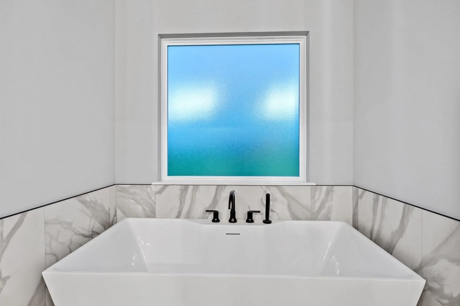 White bathtub with black fixtures in front of a large frosted window with white trim, marble tile wainscoting, gray painted walls.