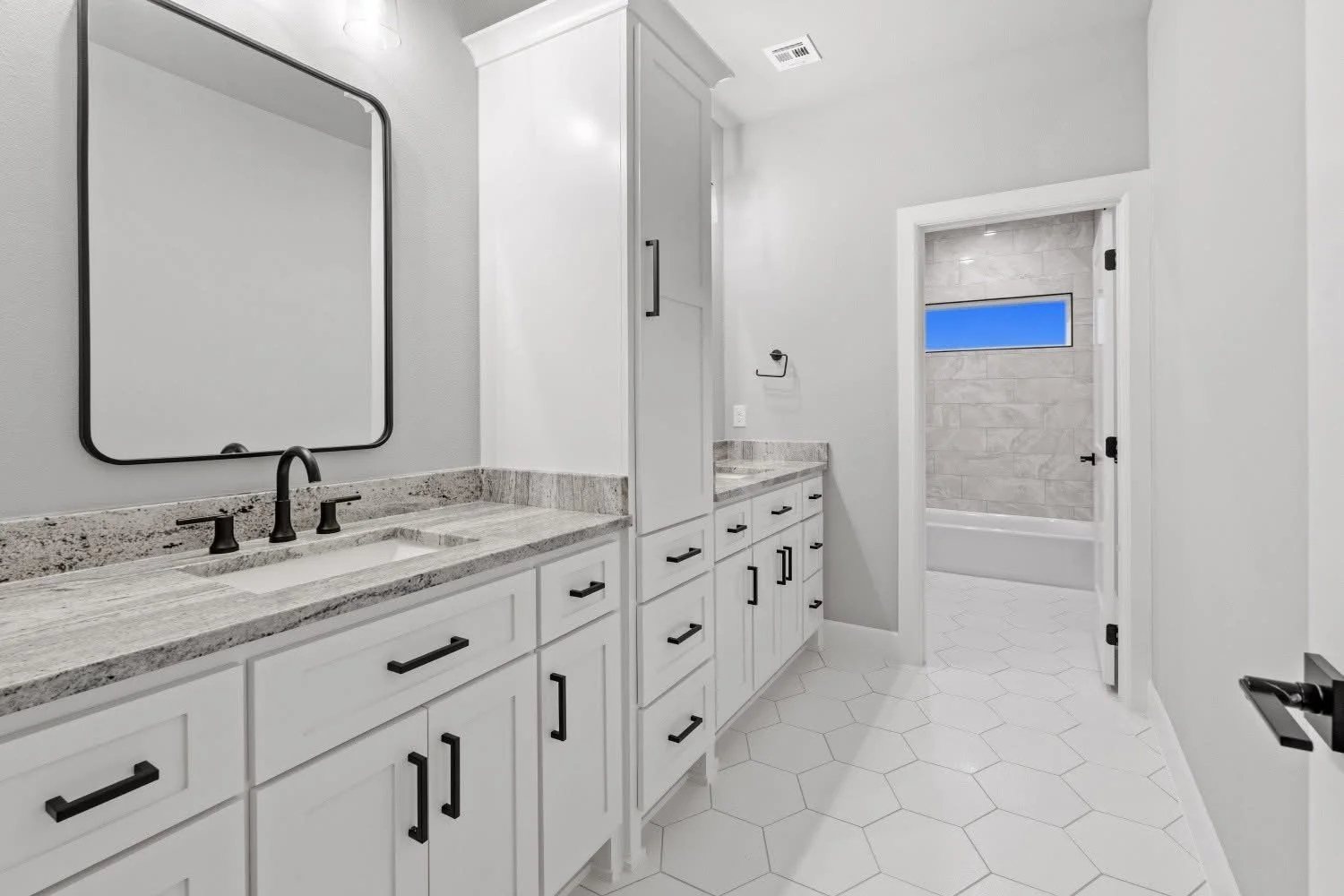Modern bathroom with white cabinets, granite countertops, black hardware, large mirror, and a shower with a horizontal window behind a door.
