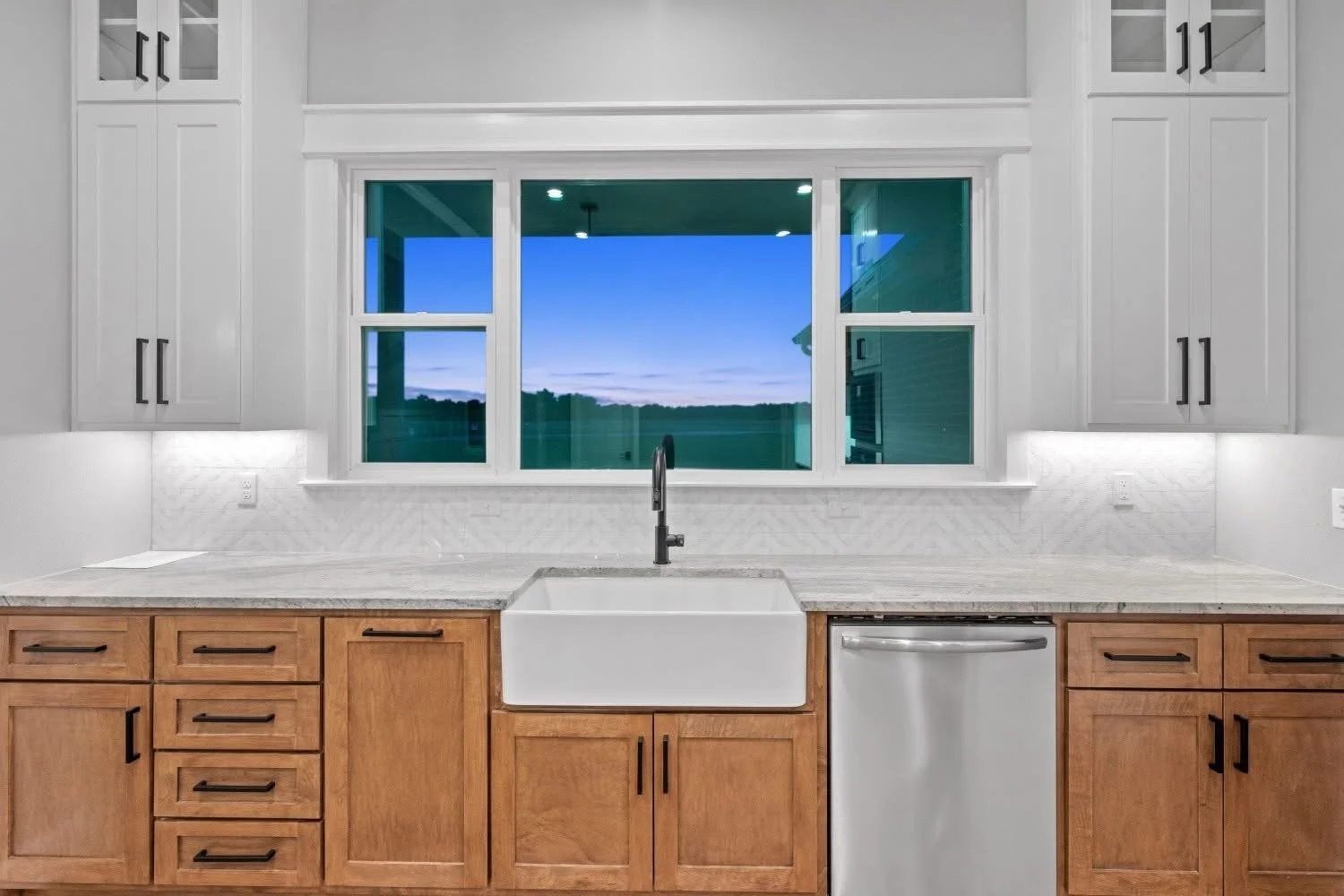 Modern kitchen with white upper cabinets, light wood lower cabinets, a white farmhouse sink, and a large window showing an evening sky with clouds.