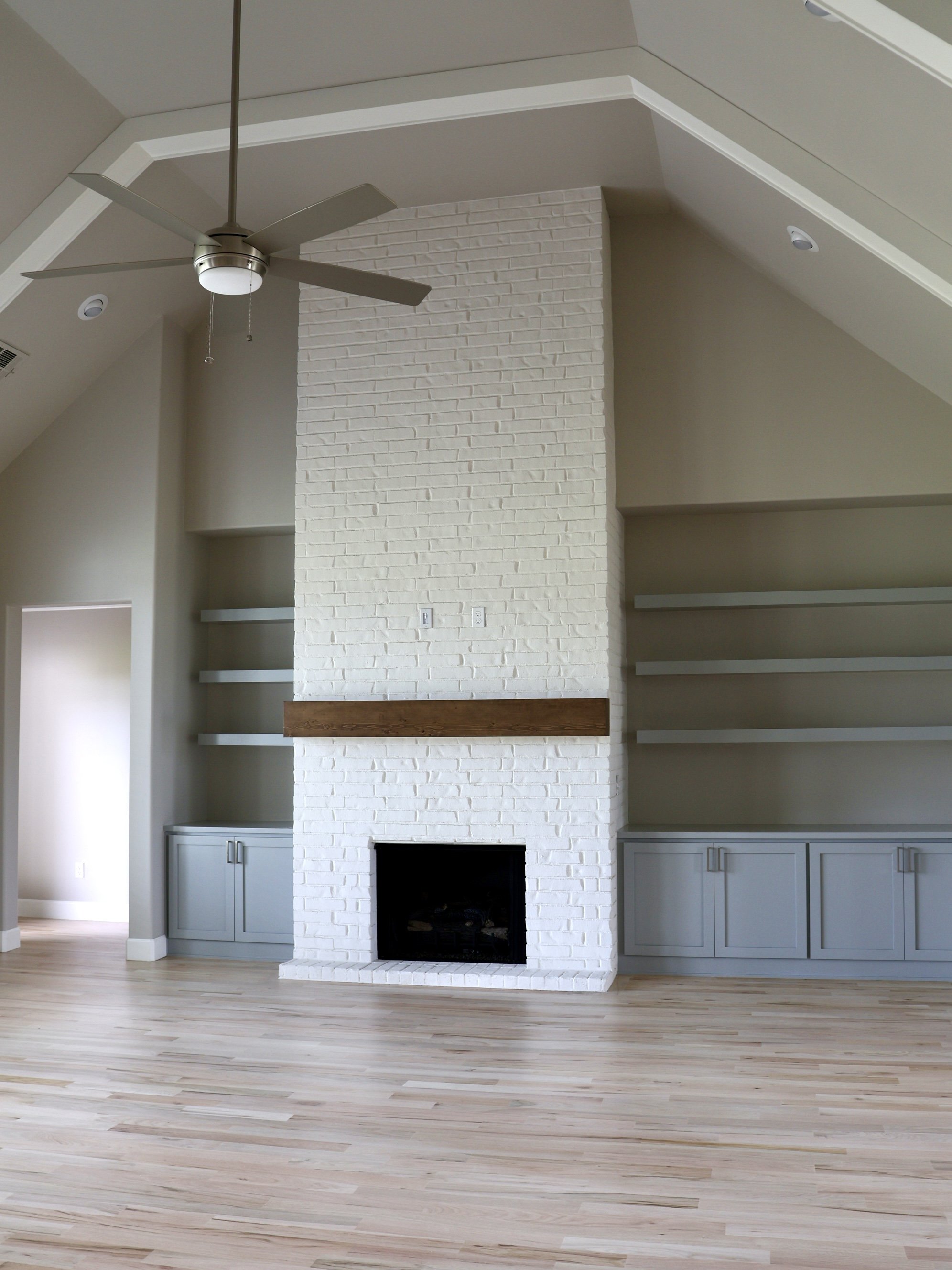 A living room with a white brick fireplace, built-in cabinets on both sides, and floating shelves above the mantel. The ceiling has a ceiling fan and recessed lighting, with light hardwood flooring.