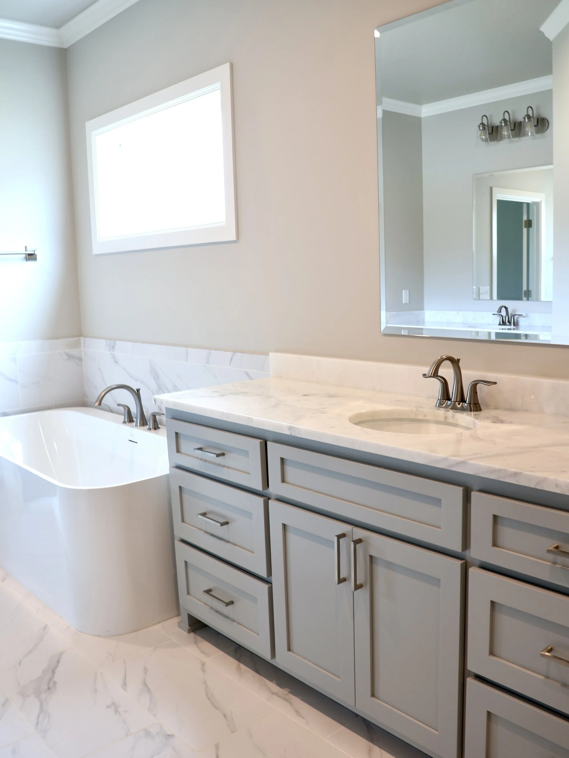 Bathroom with a marble countertop, light gray cabinets, a large mirror, a window providing natural light, and a small sink with a silver faucet.