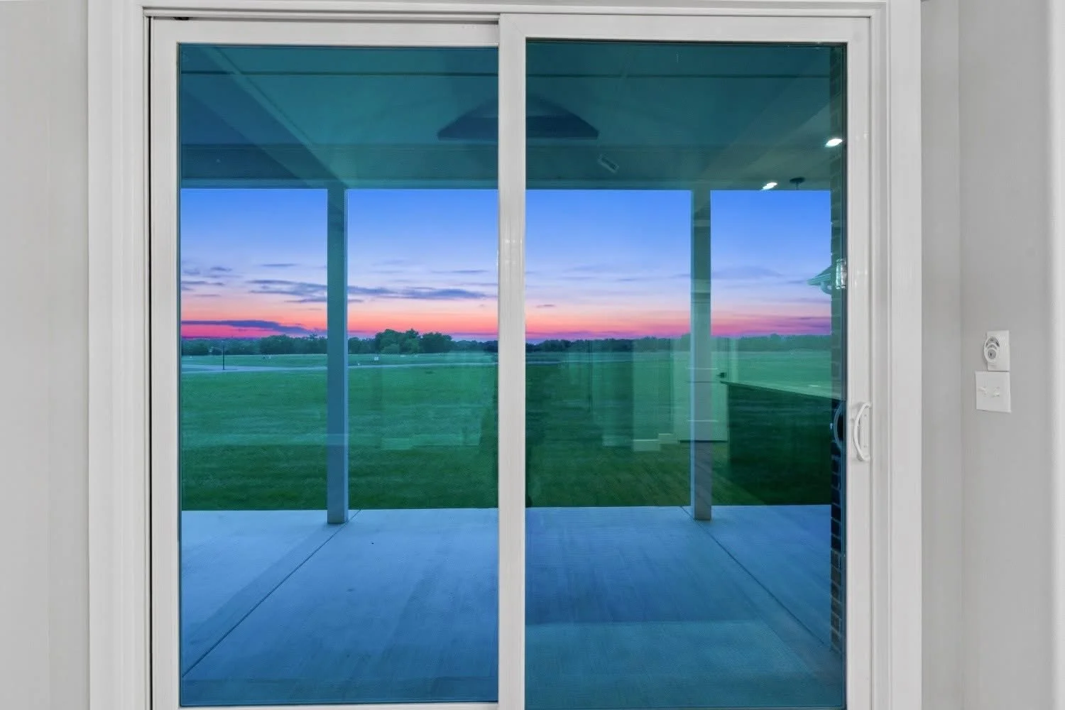 View through a sliding glass door showing a backyard with green grass and a colorful sunset sky with orange, pink, purple, and blue hues.