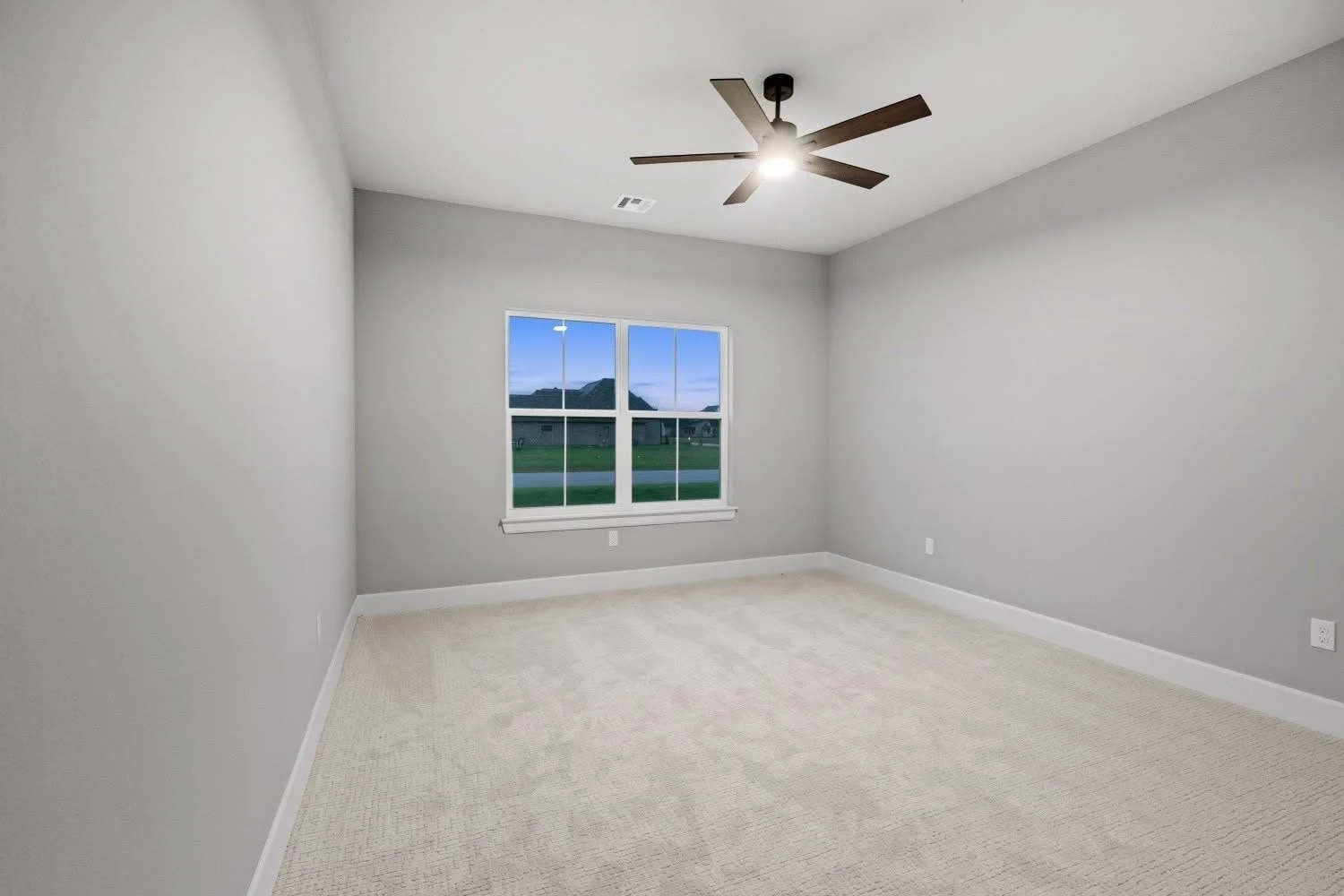 Empty room with gray walls, beige carpet, a window showing a view of neighboring houses and green lawn, and a ceiling fan with dark blades and a light.