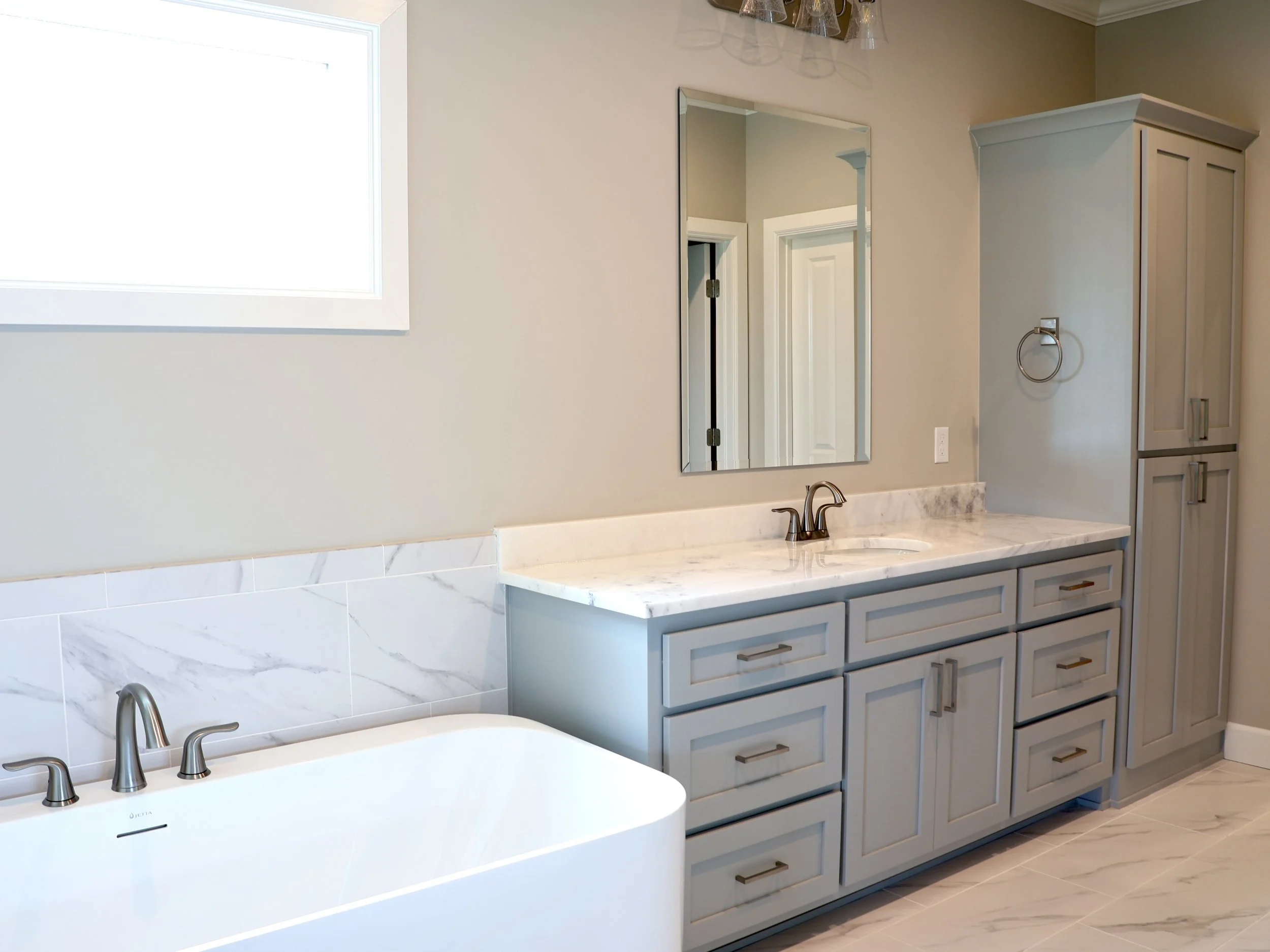 Modern bathroom with white and light gray cabinets, marble countertop, large mirror, and a bathtub with double faucets.