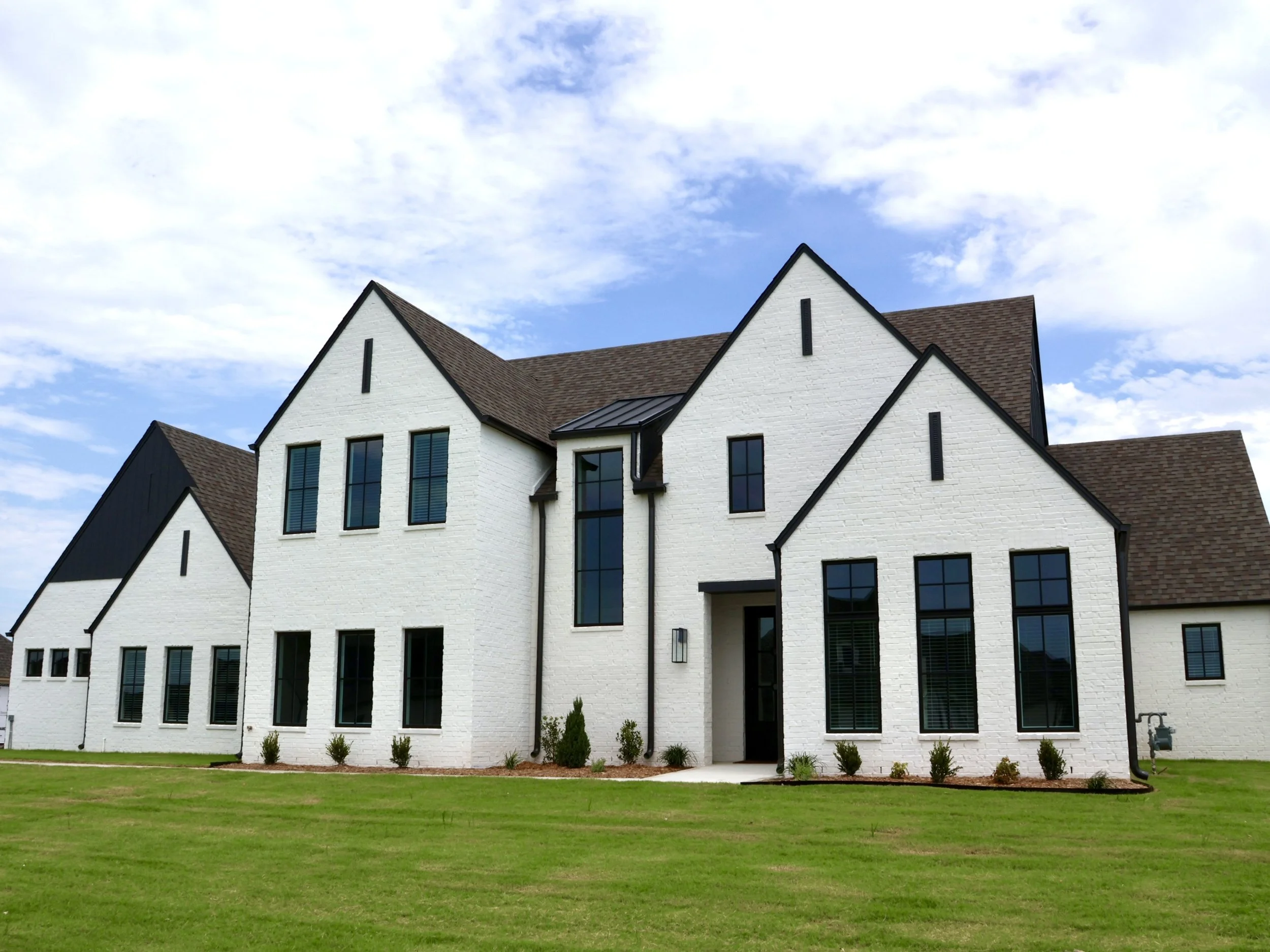 A modern white brick house with multiple peaked roofs, large windows, and black trim, situated on a well-maintained lawn under a partly cloudy sky.