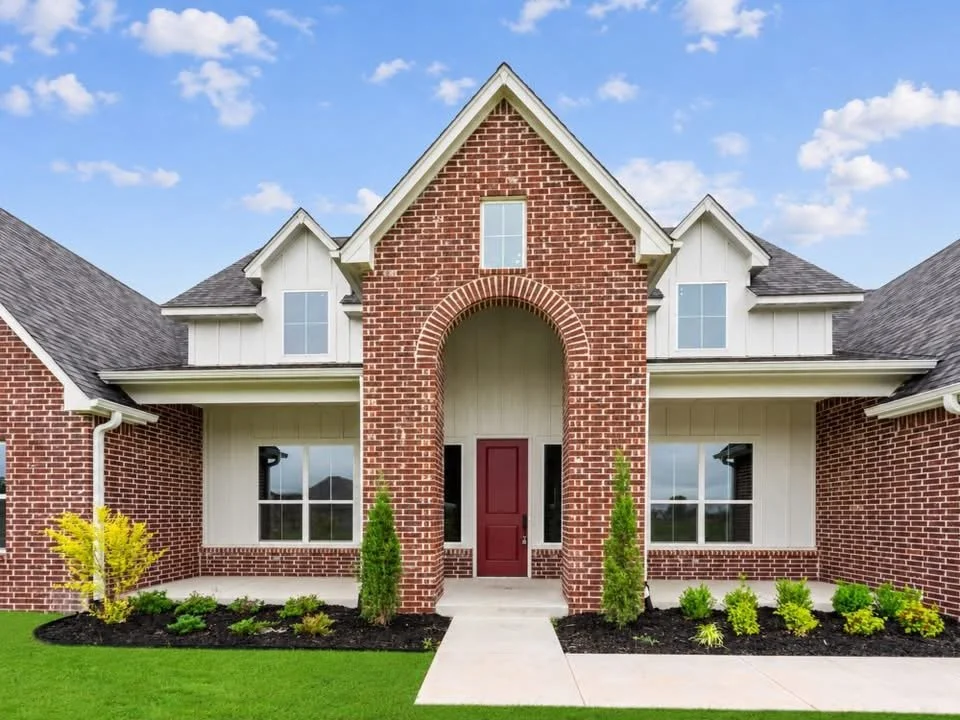 Front view of a new suburban house with red brick and white siding, a central entrance with a red door, well-maintained lawn, and blue sky.