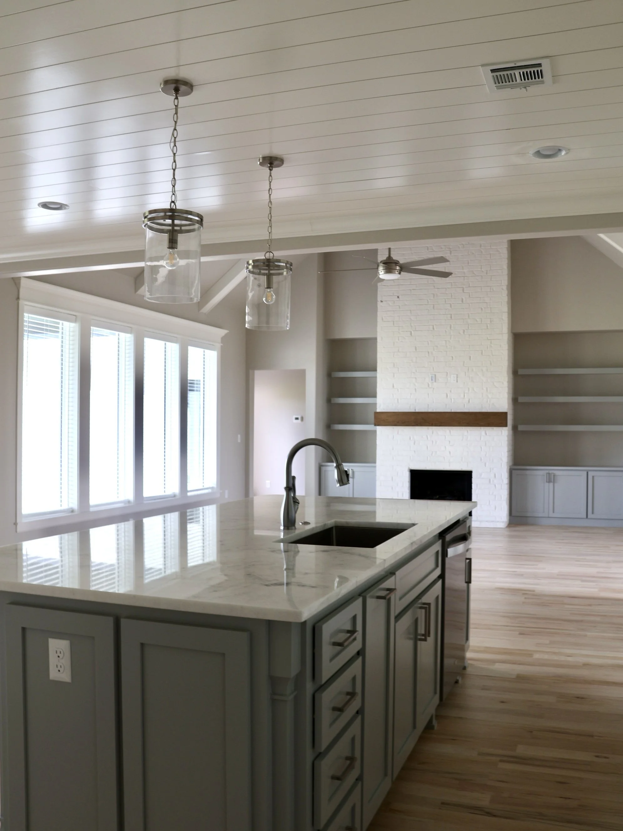 Modern kitchen with a white island, gray cabinets, large windows, a white brick fireplace, and a ceiling fan with lights.