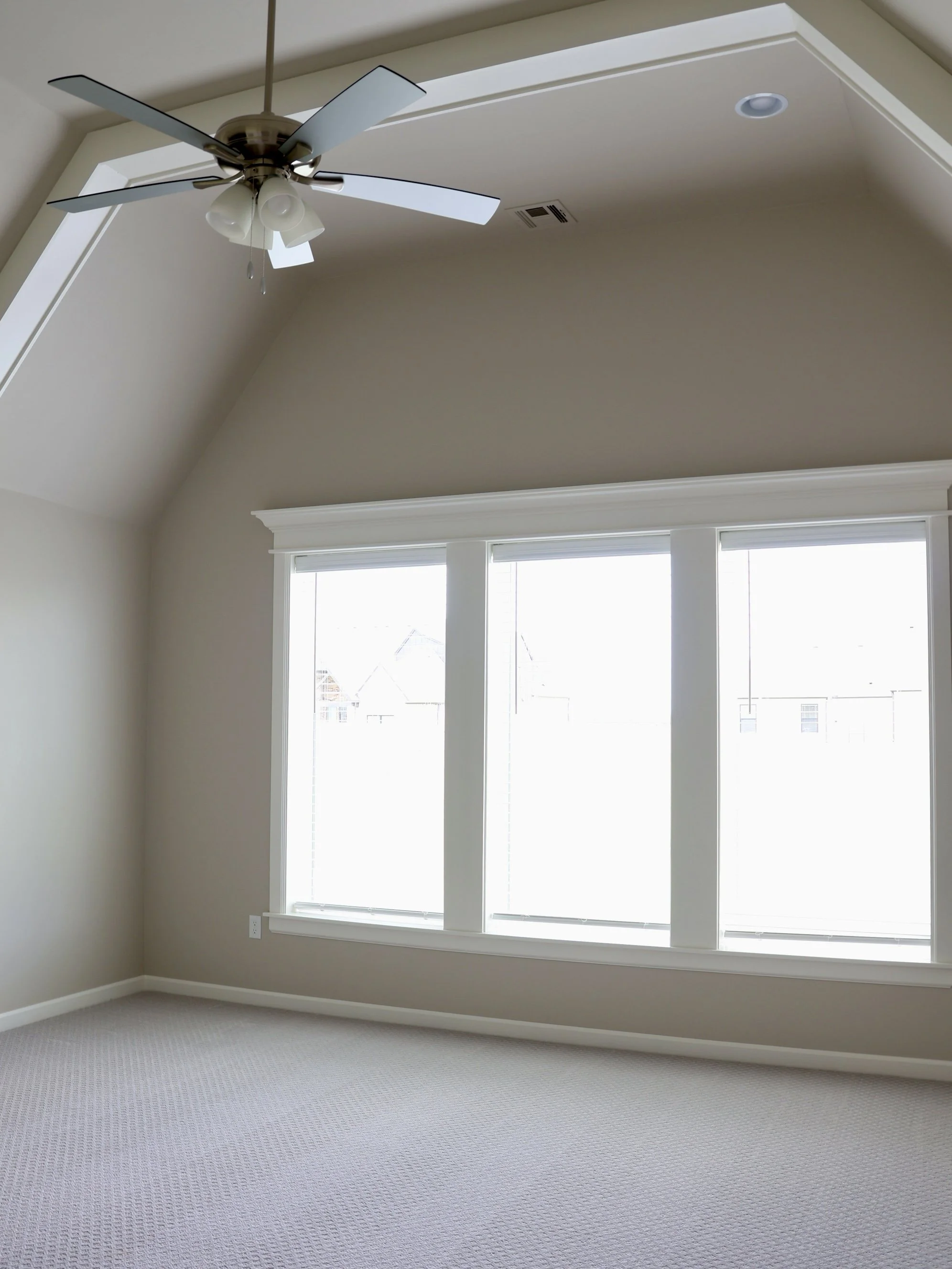 A bright, empty room with beige walls, a large window with white trim, a ceiling fan with lights, and light-colored carpeting.