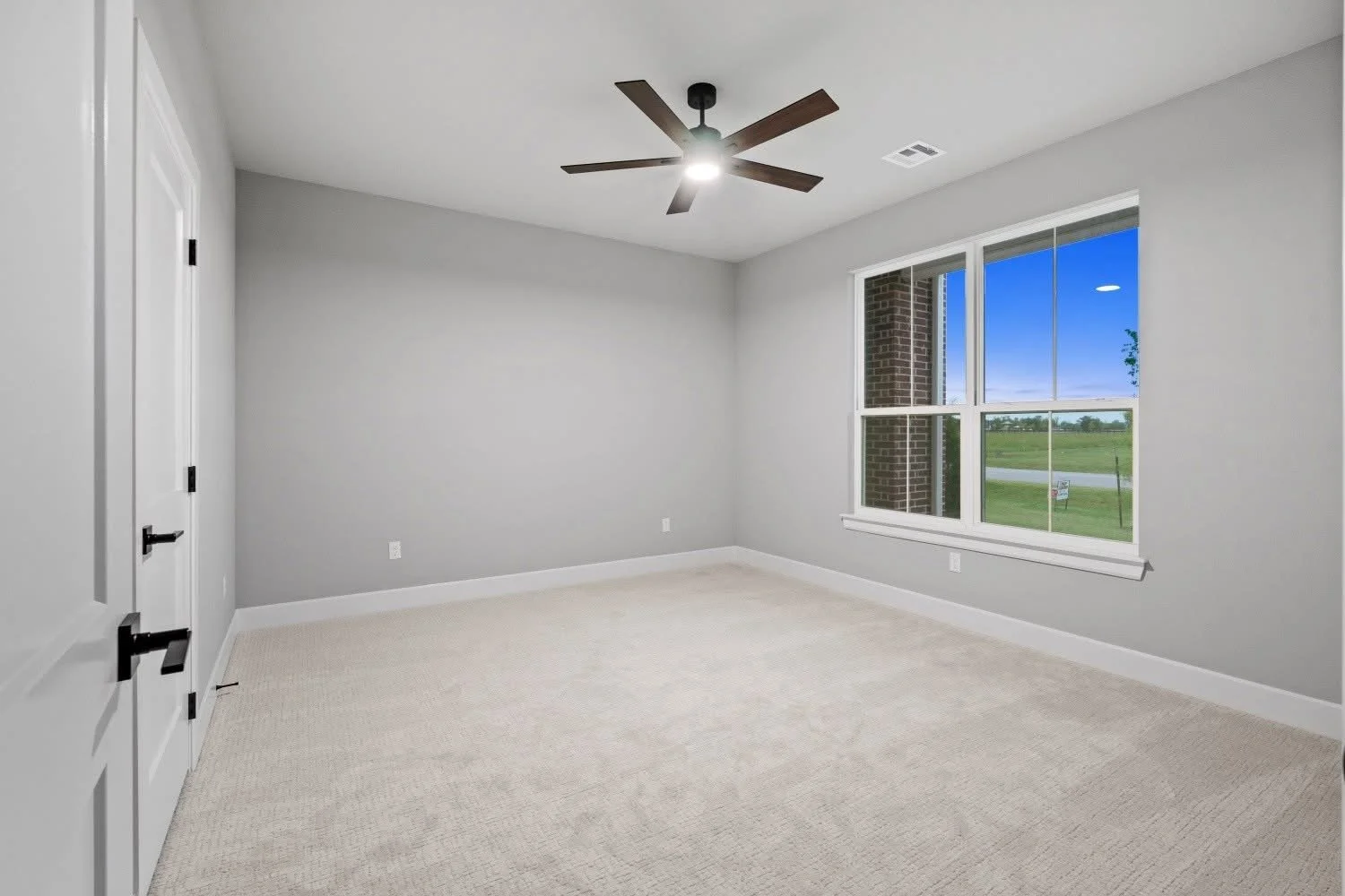 Empty bedroom with light gray walls, beige carpet, white trim, large window showing a green landscape, and a ceiling fan with wooden blades and a light.