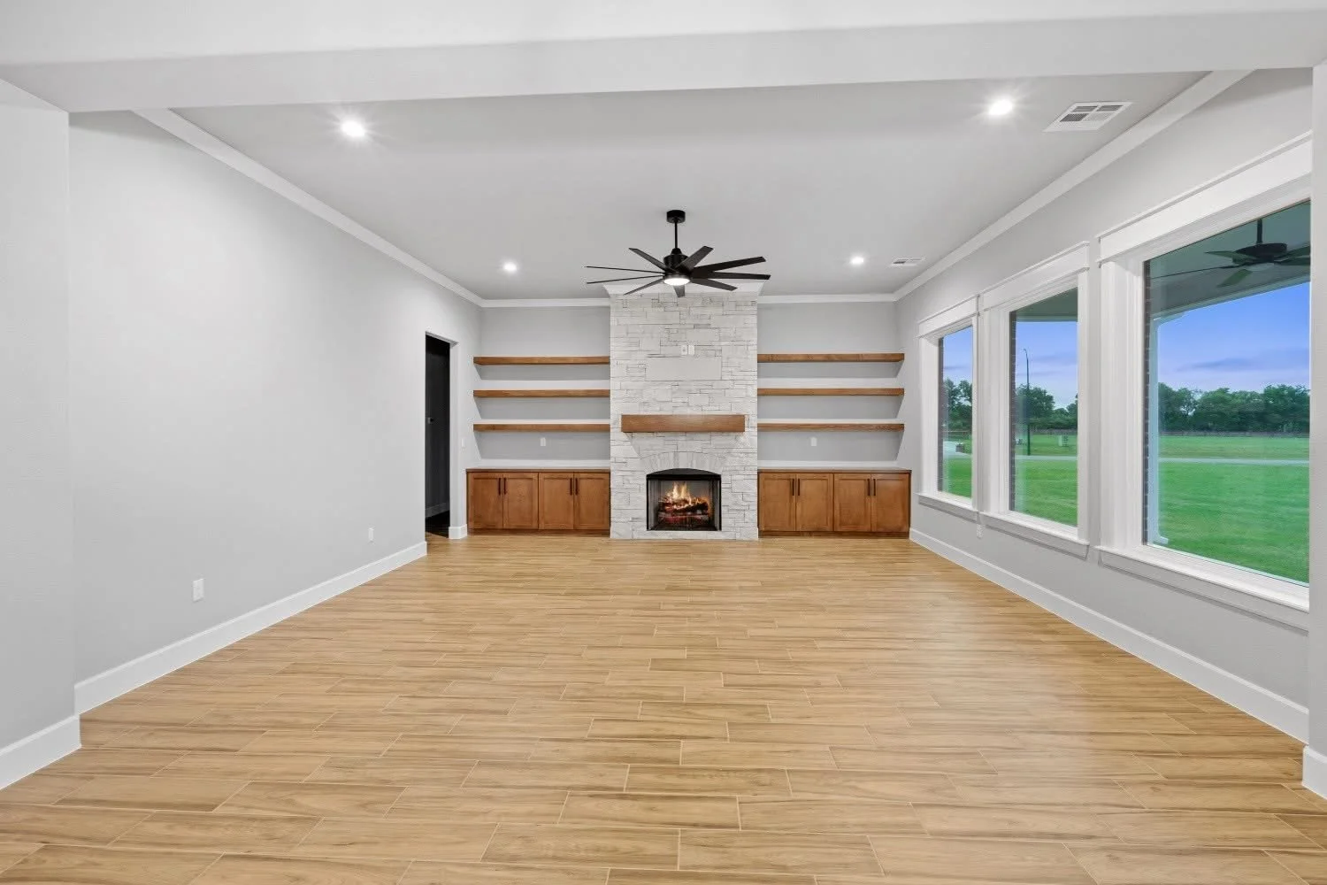 Empty living room with hardwood floors, white walls, large windows, a white brick fireplace with a wooden mantel, and built-in wooden cabinets and open shelves.