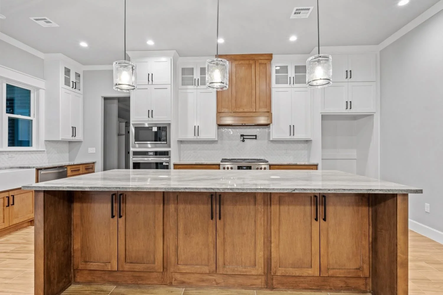 Modern kitchen with white upper cabinets, wooden lower cabinets, and a large central island with a granite countertop. Three pendant lights hang above the island. Built-in microwave, oven, stove, and a window on the left side.