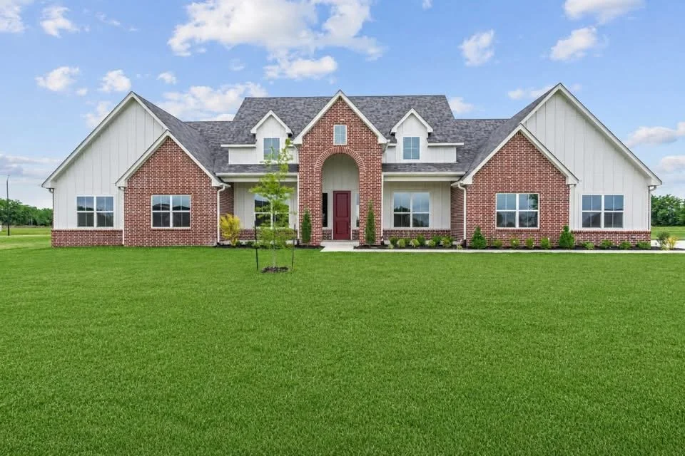 Large house with a brick and white siding exterior, gabled roof, and front yard with green grass and small trees.